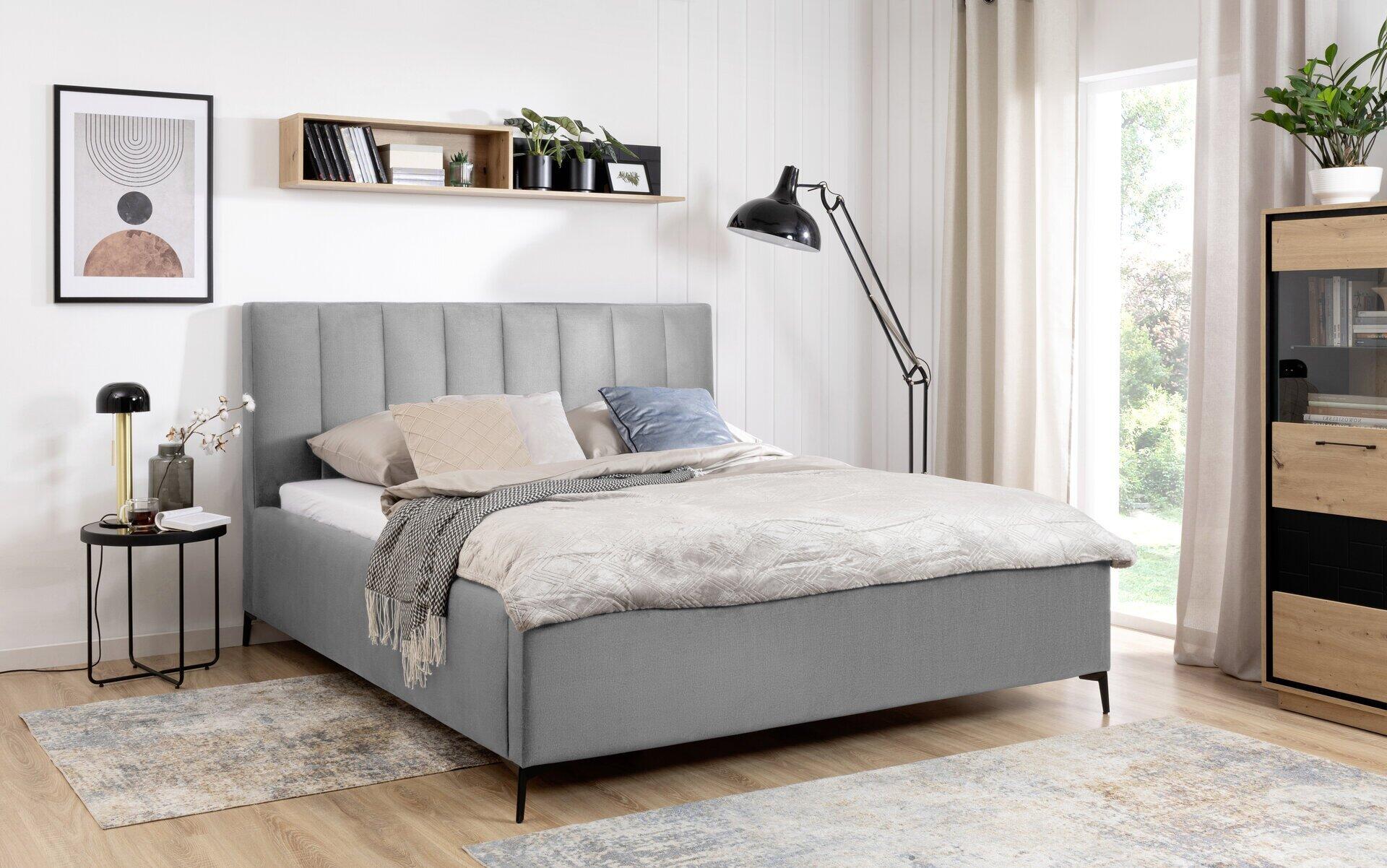 Alini Boxspringbett in hellgrau mit hohem, vertikal genähtem Kopfteil und integriertem Bettkasten, abgebildet aus leicht erhöhter, seitlicher Perspektive in einem stilvollen Schlafzimmer.