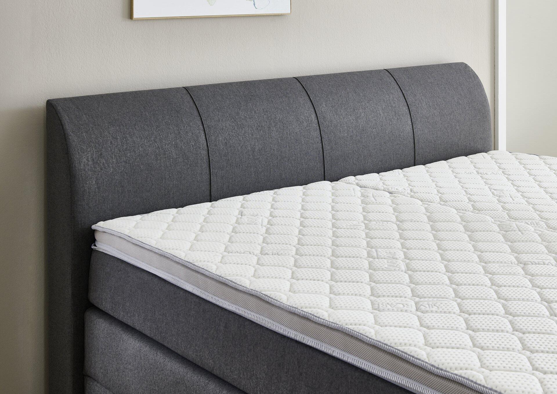 Boxspringbett Modesto BX1440 mit grauem, leicht gewölbtem Kopfteil und weißer Matratze mit cremefarbenem Topper, Detailaufnahme aus Vogelperspektive.