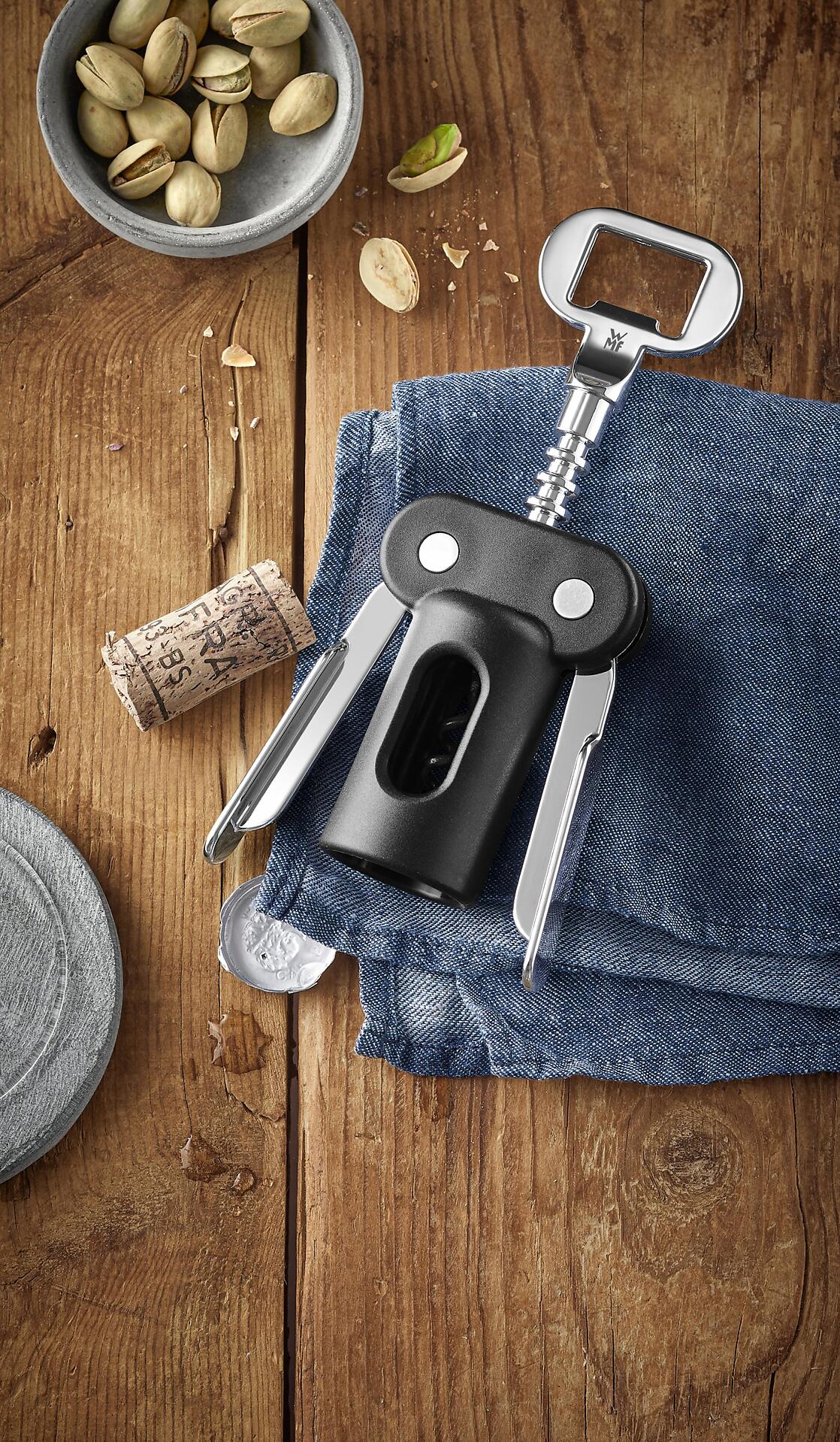 WMF Korkenzieher mit Schraubenzieher und Hebel, schräg auf Holzuntergrund vor blau-grauem Jeansstoff, mit Pistazien und Korken daneben.