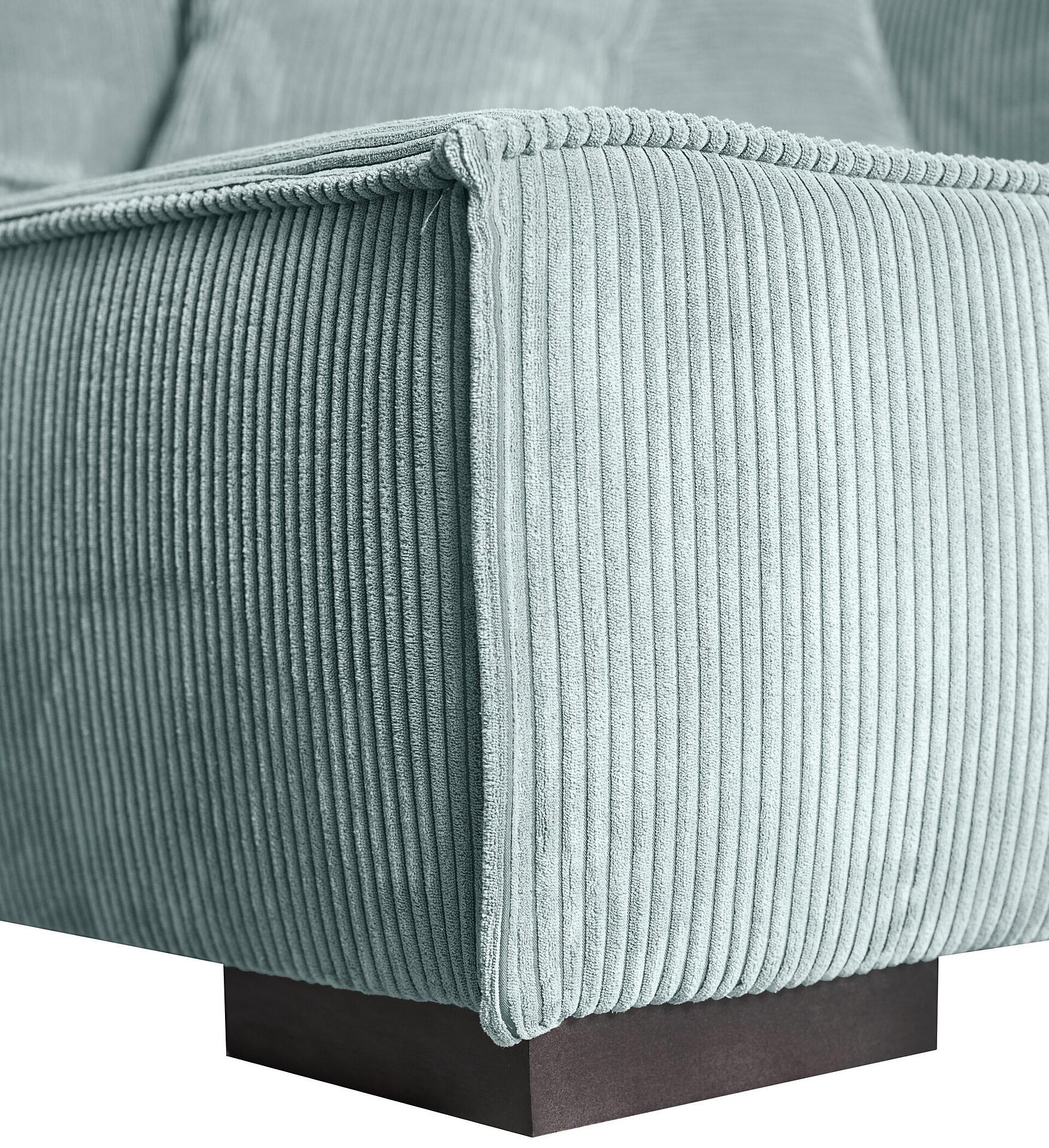 Detailaufnahme des Ecksofas Mango: Eine gerundete Armlehne trifft auf die schräg verlaufende Rückenlehne, beides mit hellgrauem Samtstoff und vertikaler Kordelstruktur, sowie dunklen, kubischen Holzfüßen.