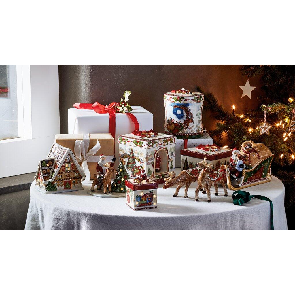Festlich dekorierte Weihnachtsdorf-Szene mit Lebkuchenhaus mit Spieluhr, Chalet, beleuchteten Häusern, Figuren und Geschenken vor einem geschmückten Weihnachtsbaum.