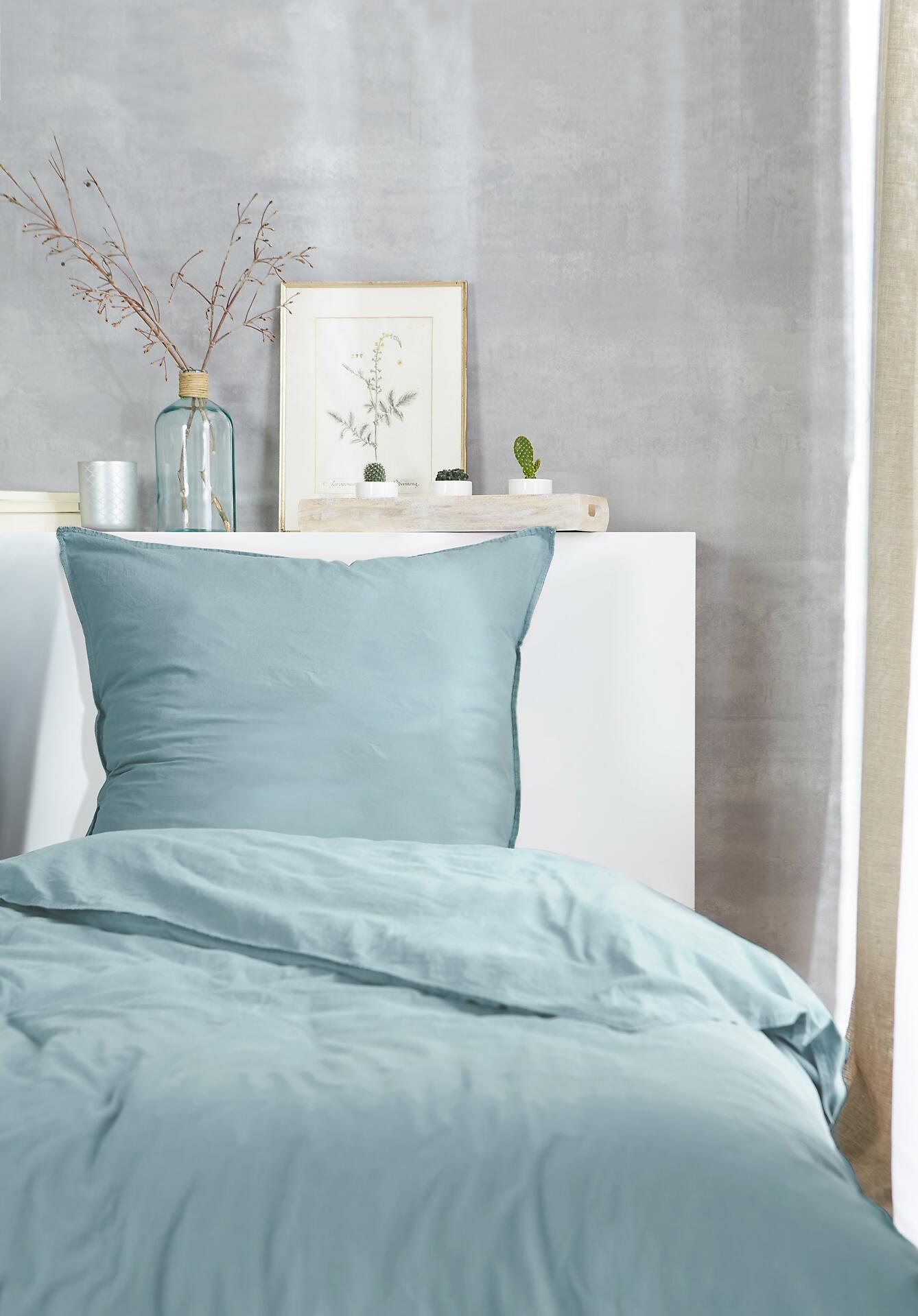Nahaufnahme einer graugrünen, Stone-Washed Renforcé Bettwäsche aus Baumwolle auf einem blauen Bett, mit Reißverschluss und 80x80cm Kissen.