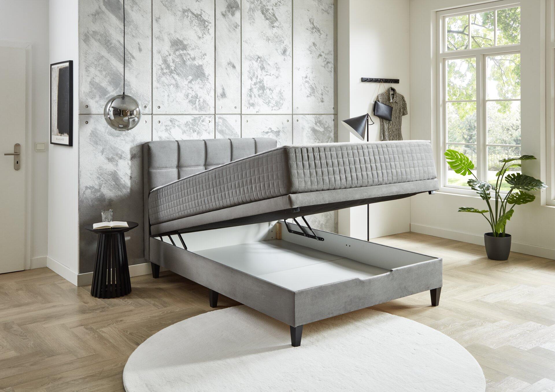 Boxspringbett Wien BX2590 mit grauem Samtbezug und hochklappbarem Bettgestell, das Stauraum freigibt, aus leichter Seitenperspektive fotografiert.