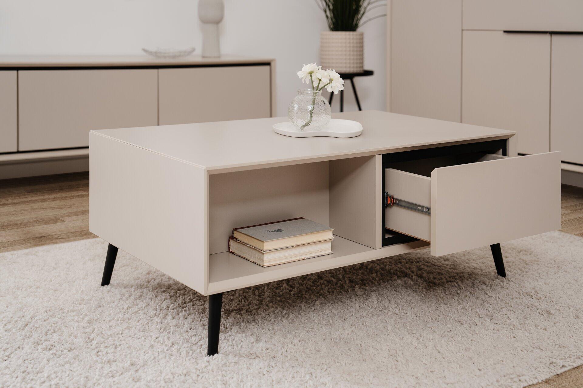 Couchtisch Arona 55A mit rechteckiger Form, hellem beige-grauem Korpus, einer Schublade links, offenem Fach mit Büchern rechts und schlanken, schwarzen Tischbeinen.