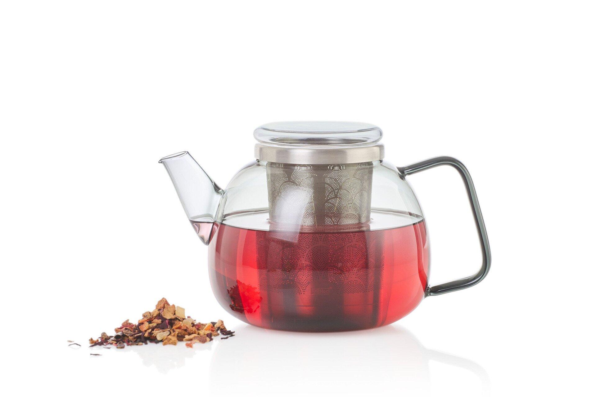 Teekanne Yuna mit transparentem Glas und Edelstahl-Siebdeckel, gefüllt mit rotem Tee und neben Hibiskusblüten.