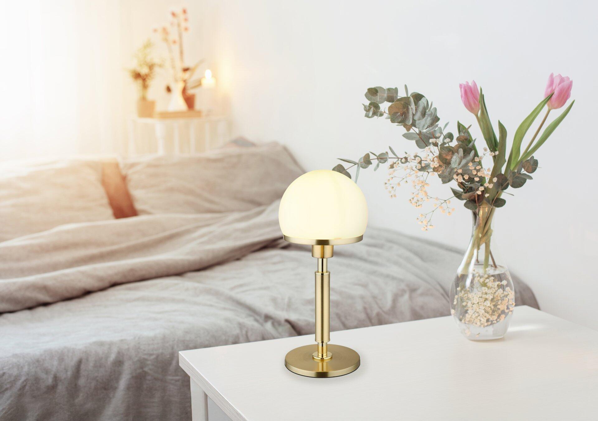 Goldfarbene Tischleuchte HAKU mit mattiertem, rundem Lampenschirm und Wippschalter am Kabel, stilvoll arrangiert auf einer Kommode mit Blumenvase.