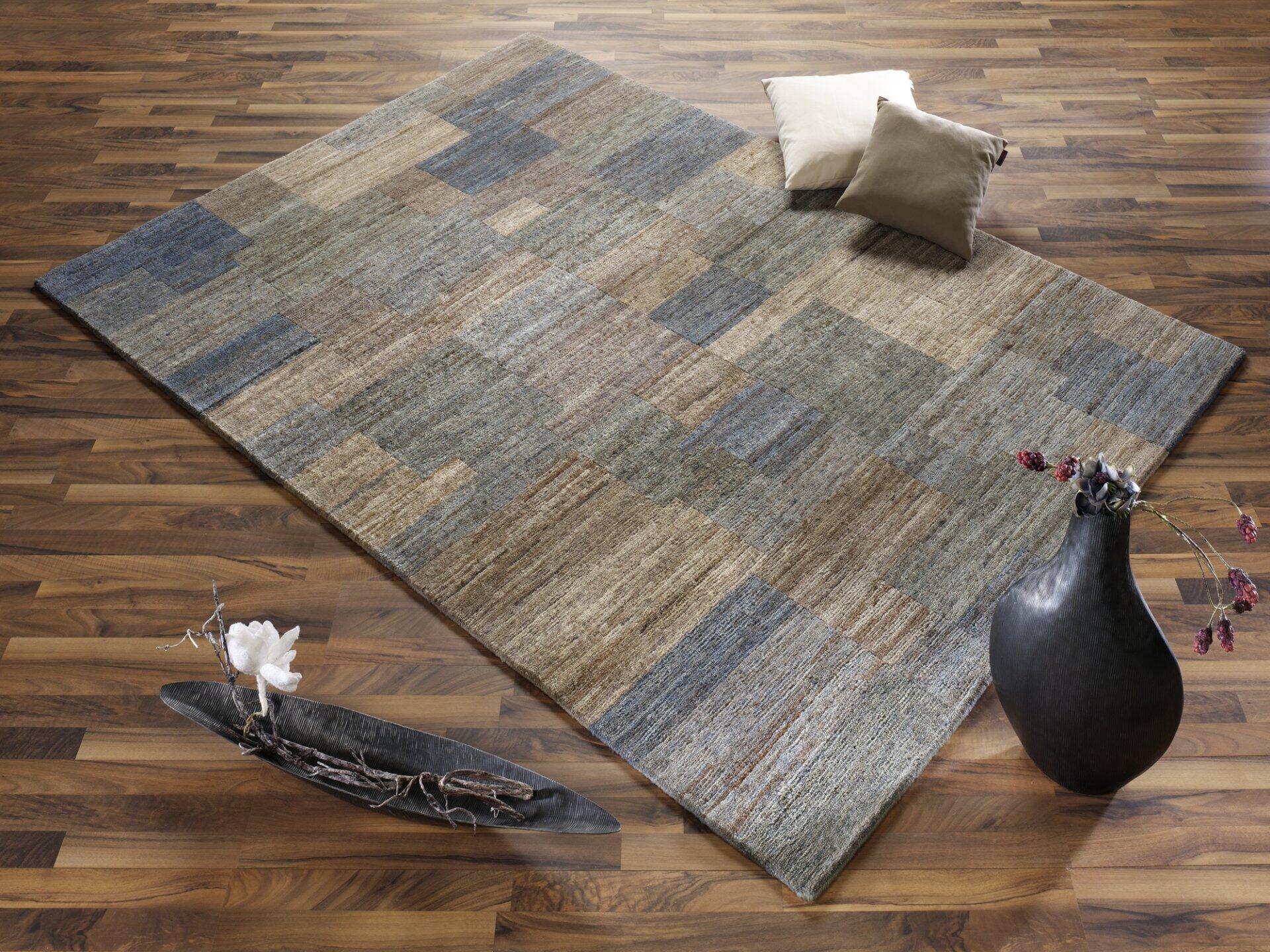 Handknüpfer Teppich Badipur mit horizontalen Streifen in Grau- und Beigetönen, diagonal auf einem Holzboden in einem modernen Wohnambiente.