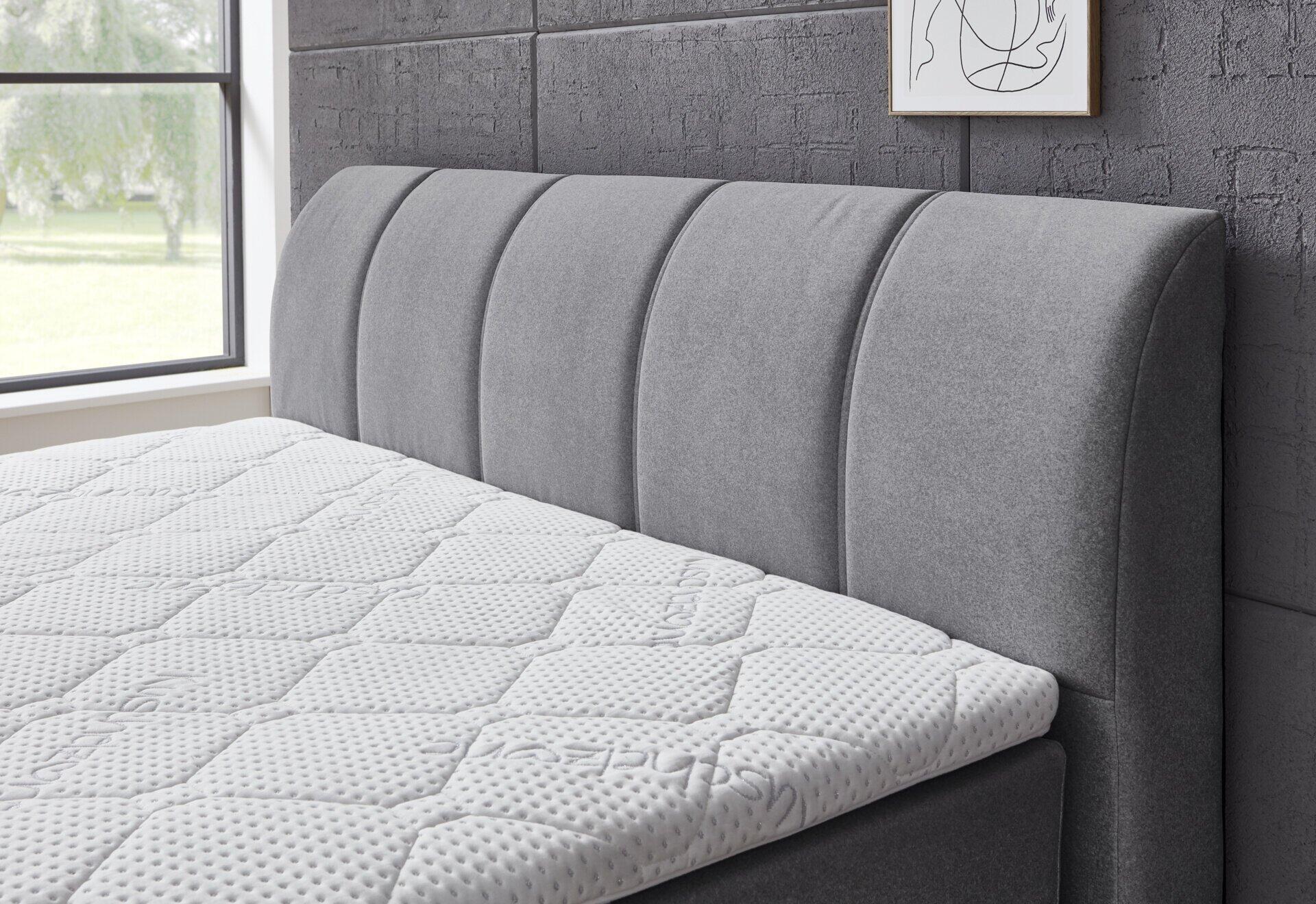 Boxspringbett Graz BX2630 mit grauem Bezug, hochgepolstertem Kopfbereich mit vertikalen Fugen und integrierter Stauraumlösung, aus leicht erhöhter Perspektive.