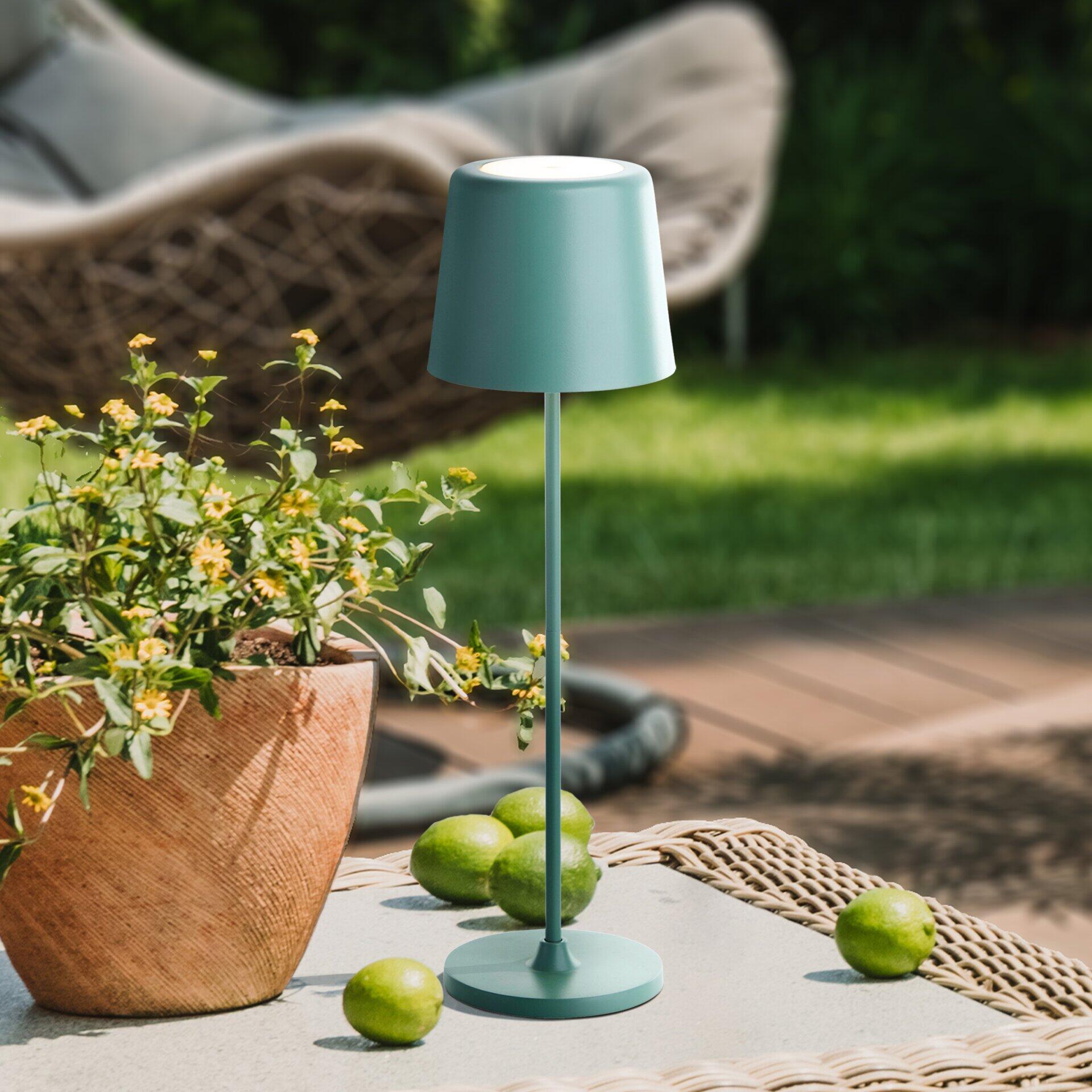 Design-Tischleuchte Kaami im Grünton mit zylindrischem Lampenschirm und Stabfuß auf einem runden Sockel, detailreich dargestellt auf einer Rattan-Liege.