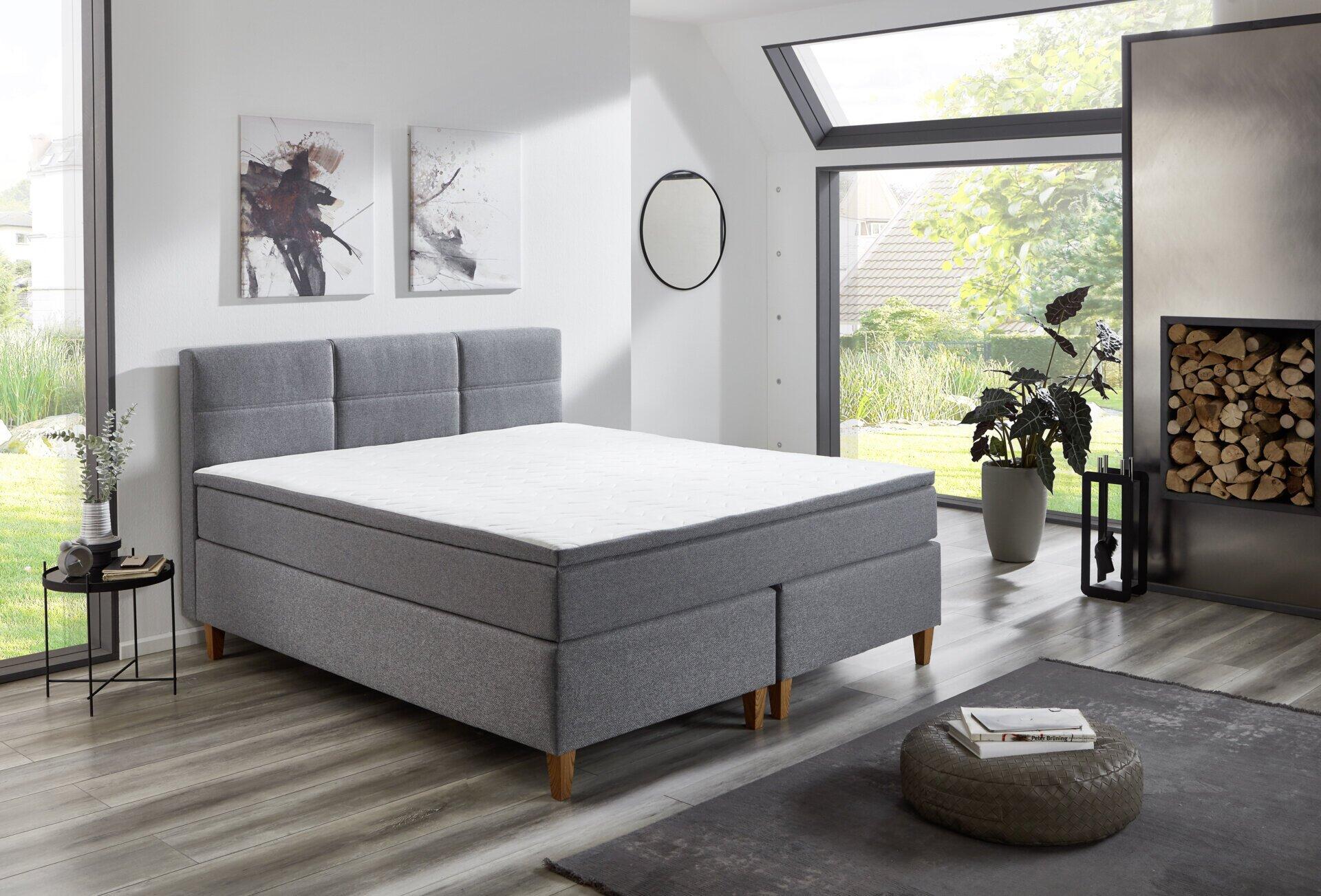 Boxspringbett Kenora BX2360 mit grauem Stoffbezug, hellem Holzfuß, integriertem Stauraum und einer weißen Matratze, frontal fotografiert im modernen Schlafzimmer.