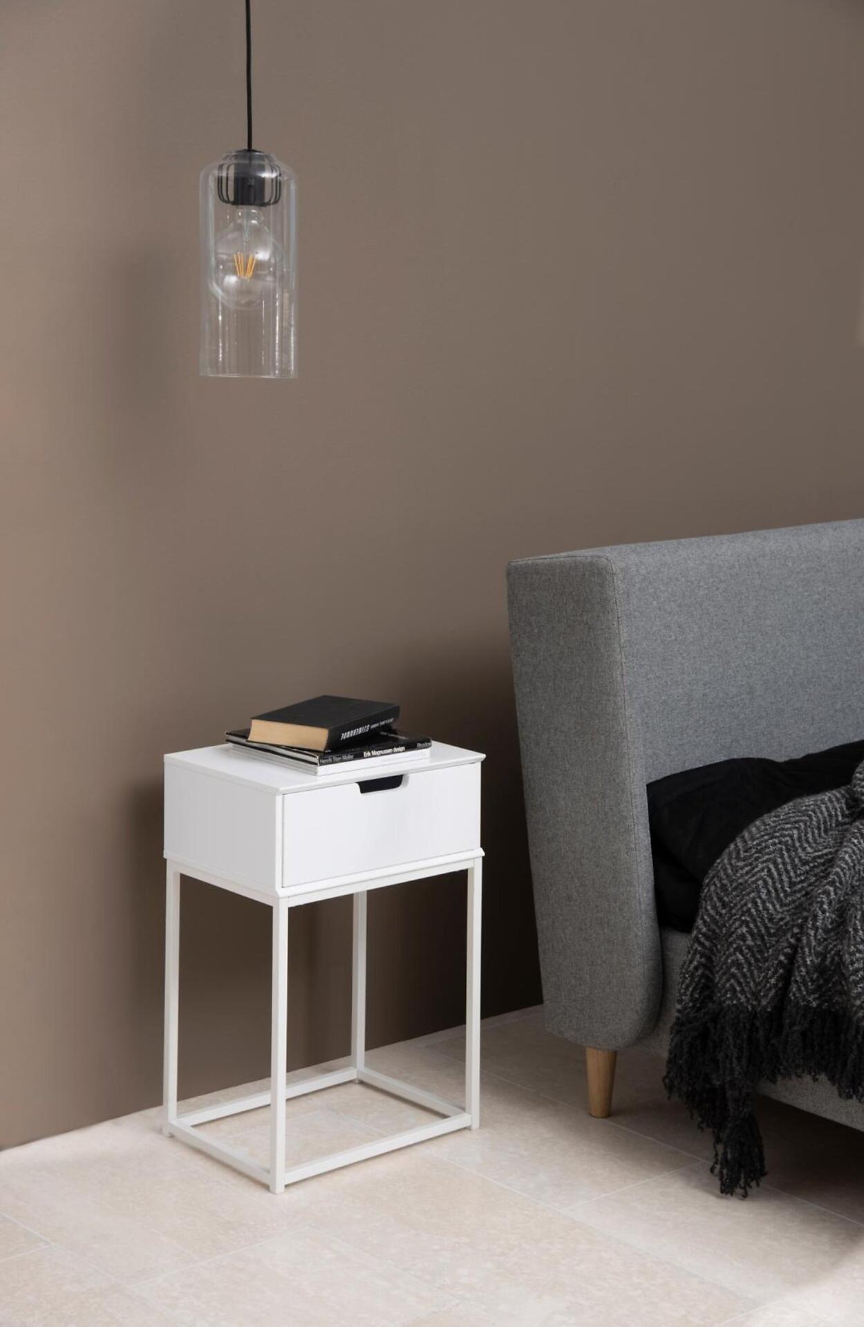 Nachttisch Mitra in Weiß mit einer Schublade und filigranen Metallbeinen, gezeigt aus schräger Perspektive mit Buch und Notizblock auf der Tischplatte.