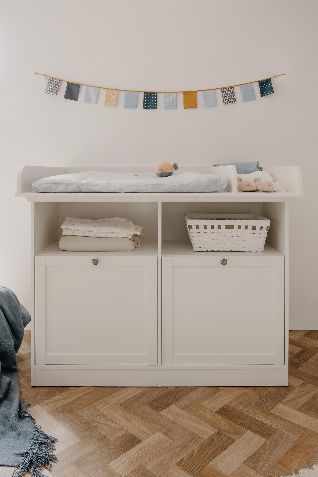 Weiße Wickelkommode Landwood 67A mit abnehmbarem Wickelaufsatz, zwei Türen mit antik-Optik-griffen und zwei Schubladen, sowie offenen Fächern für Windeln und Zubehör.