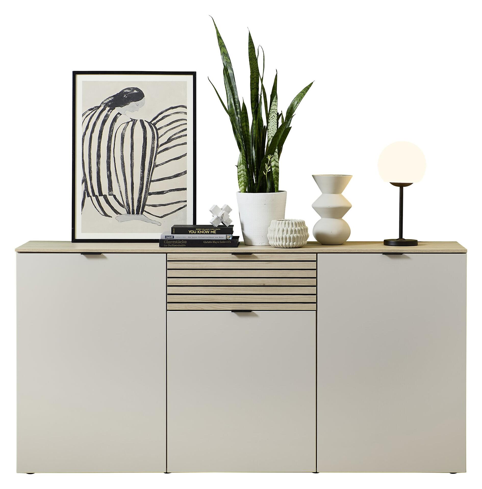 Weißes Sideboard Ina mit Riva Eiche Nb-Optik, drei Schubladen und einer Tür, dargestellt in einer leicht erhöhten Frontalperspektive in stilisierten Wohnumgebung.
