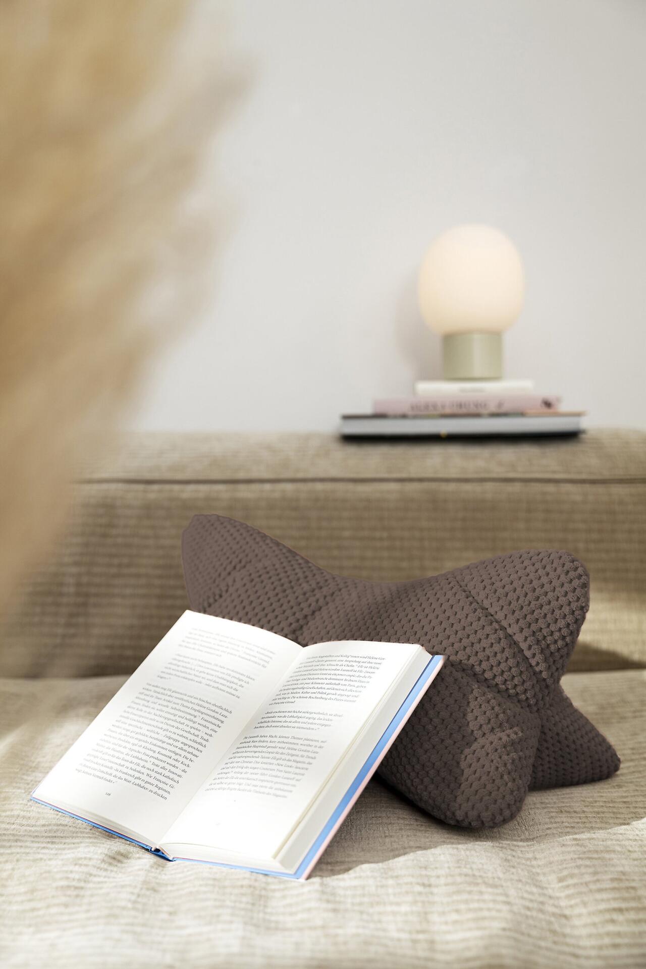 Lesekissen Comodo in Herzform auf einem Sofa mit einem aufgeschlagenen Buch, das die Funktion als bequeme Büchstütze zeigt.