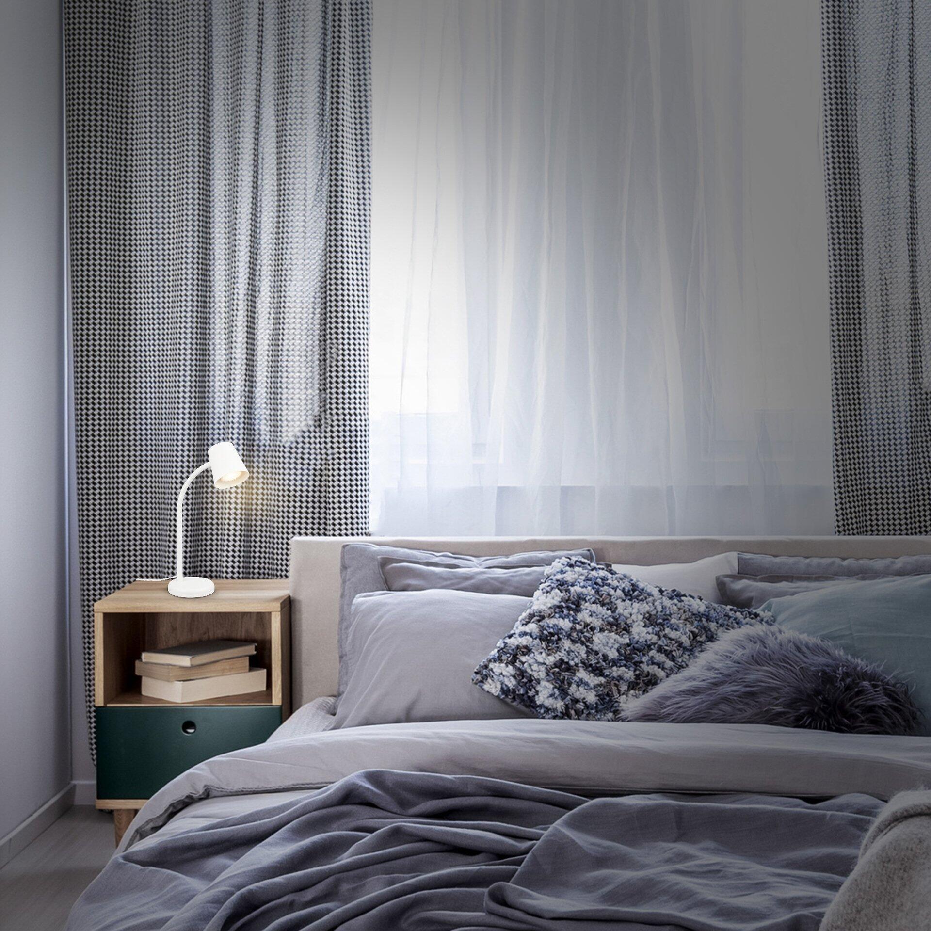Tischleuchte Skriva mit LED-Beleuchtung auf einem Nachttisch mit Schublade und offener Ablagefläche vor einem Bett.
