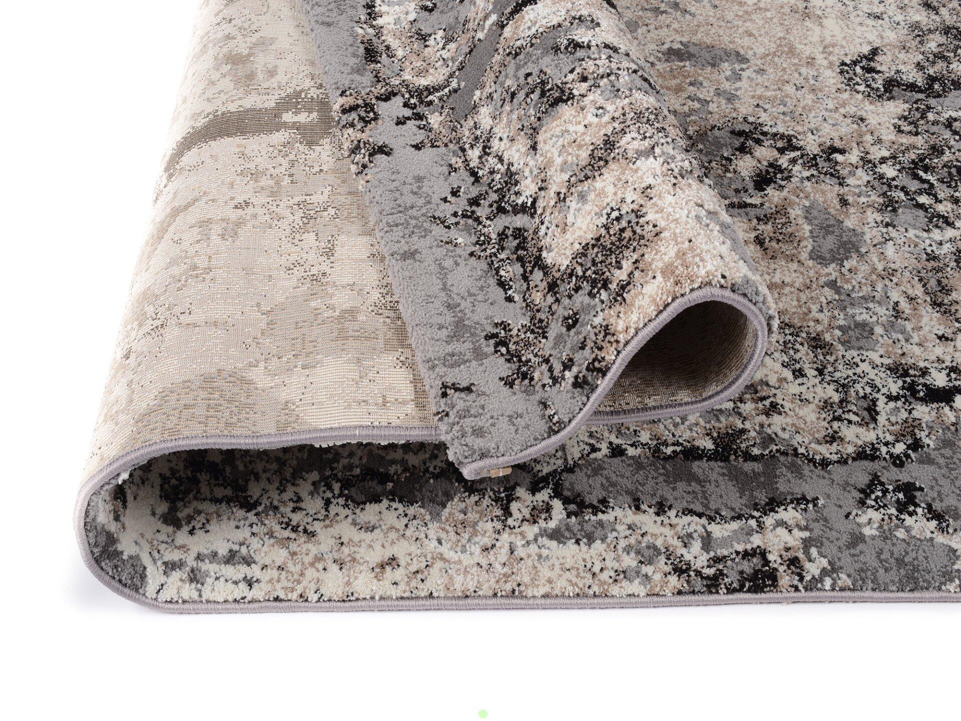 Maschinenwebteppich Koralka, detailreiche Aufnahme aus schräger Vogelperspektive, zeigt gefalteten Teppich mit gemustertem Oberflächen-Design in Grau- und Beigetönen sowie hellgraue Unterseite mit verstärktem Rand.