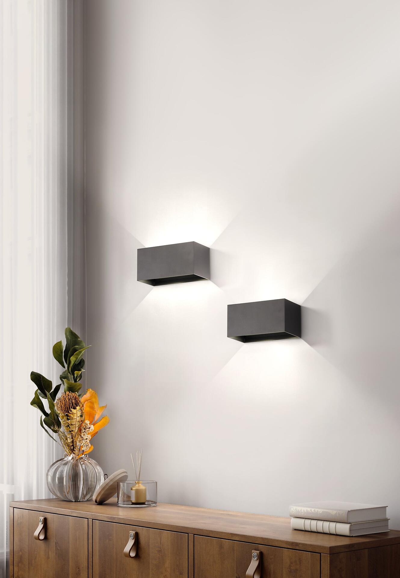 Zwei quadratische Wandleuchten Wallo mit warmweißer LED-Beleuchtung, an einer hellen Wand in unterschiedlicher Höhe angebracht, ergänzt durch ein Möbelstück mit Ledergriffen.
