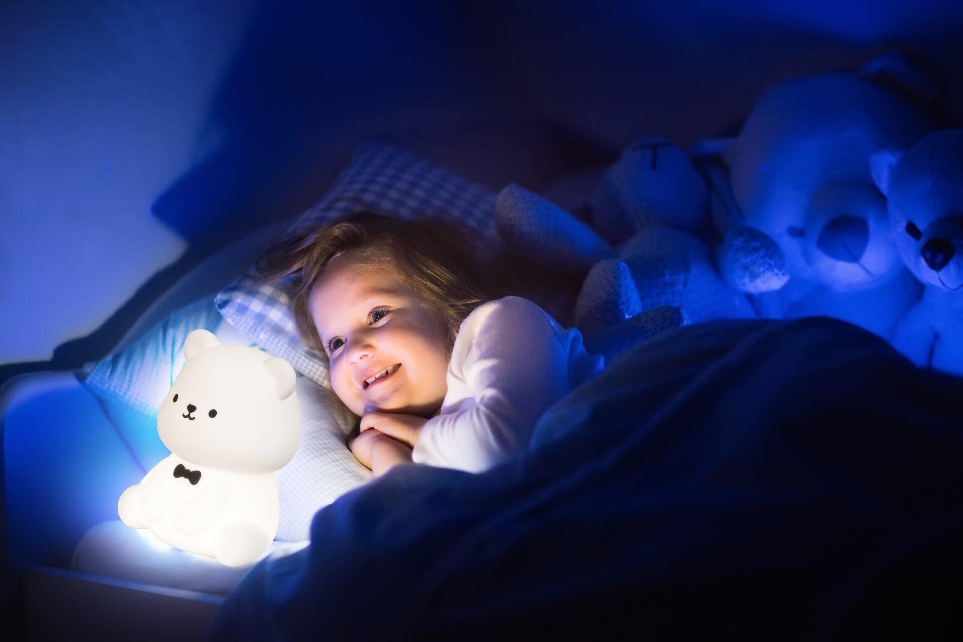 Tischleuchte Karl als Teddybärenlampe mit freundlichem Gesicht und Fliege, die im Dunkeln ein blaues Licht auf ein Kind im Bett wirft.