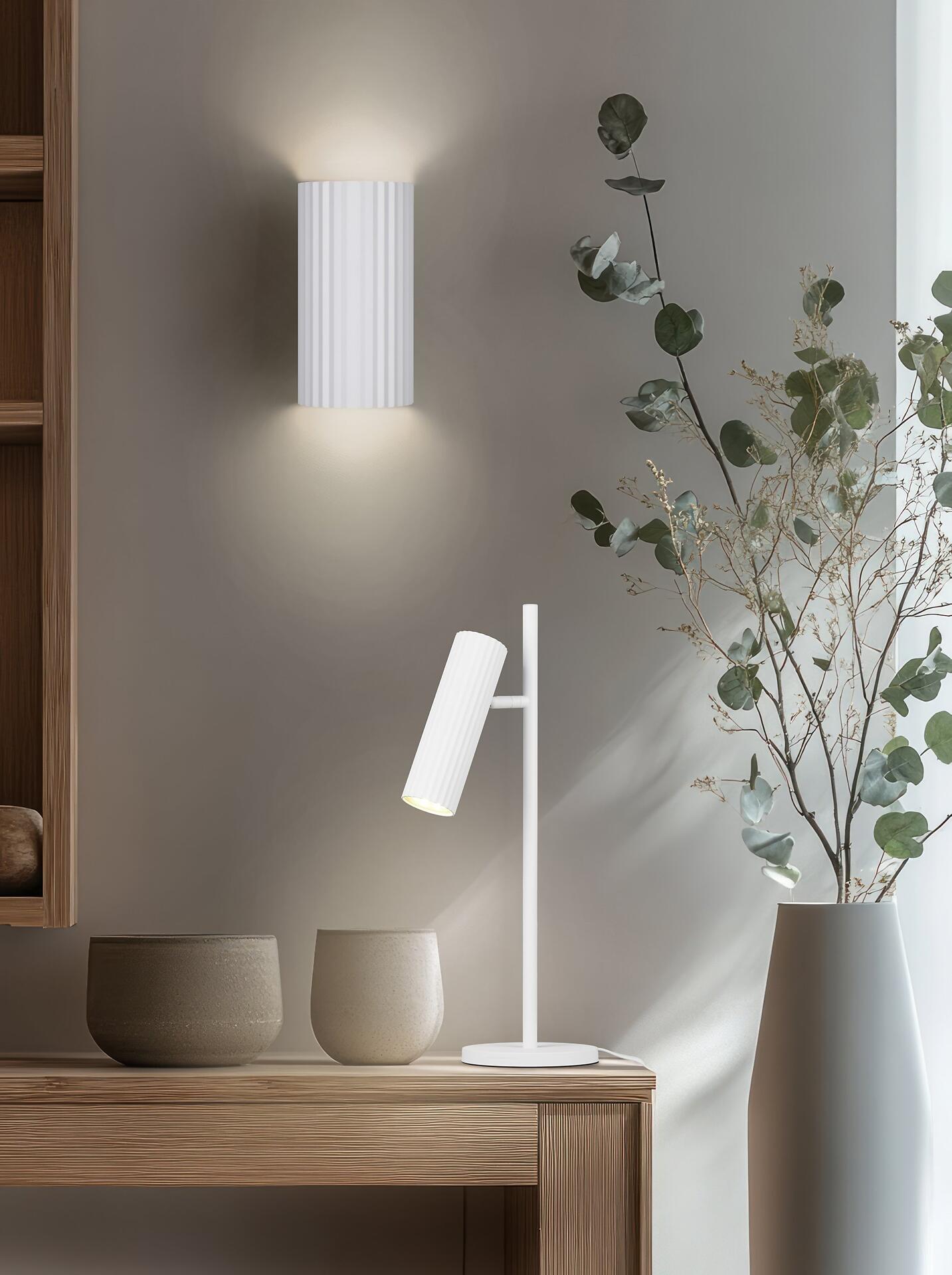 Wandleuchte Soka mit zylindrischem Lampenschirm, die in leicht erhöhter Perspektive vor einer hellen Wand einen hellen, aufwärts gerichteten Lichtschein erzeugt und die flexible Ausrichtung des beweglichen Lampenkopfs zeigt.