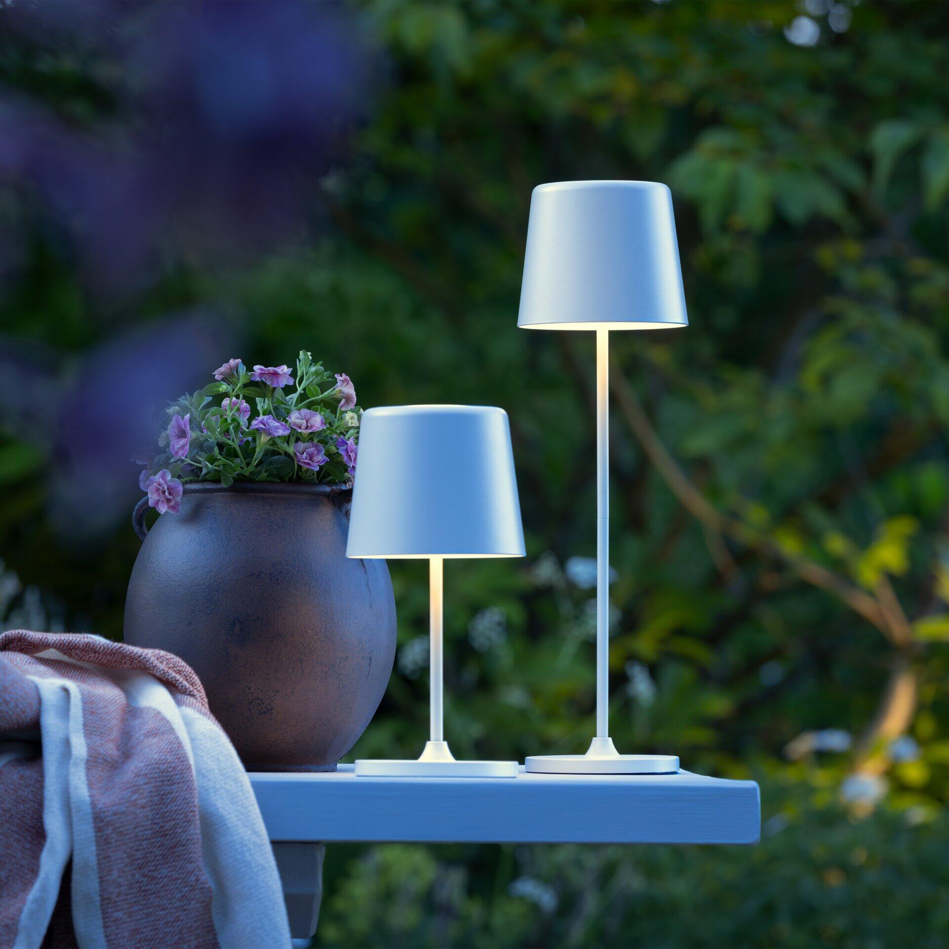 Zwei batteriebetriebene Tischlampen des Typs Kaami beleuchten im Dämmerlicht eine Terrasse und zeigen ihre kabellose Flexibilität und schlanke, leicht geneigte Form aus einer leicht erhöhten Perspektive.
