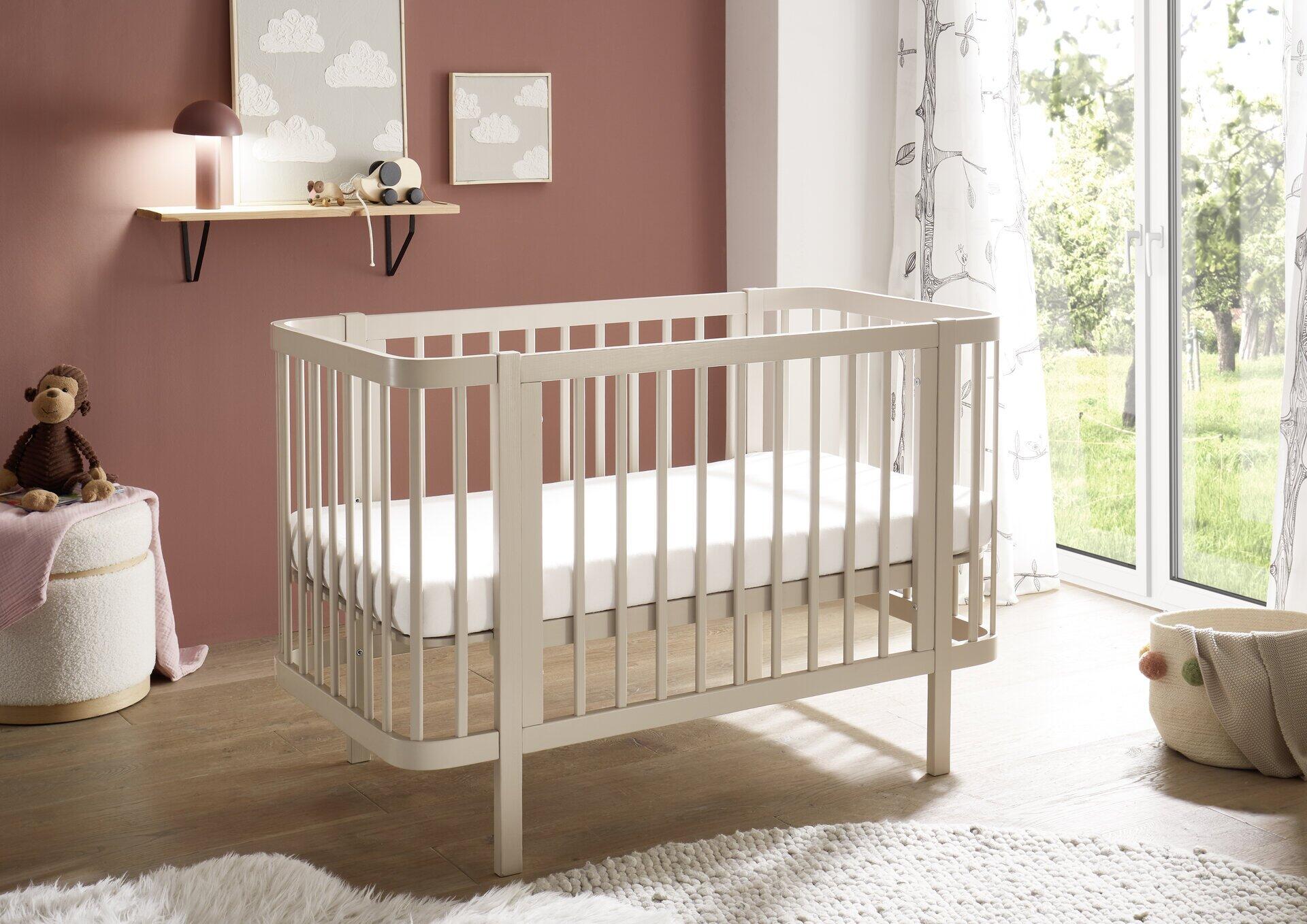 Babybett Peppini im ¾-Format mit abgerundeten Ecken und Gitterstäben vor roter Wand.