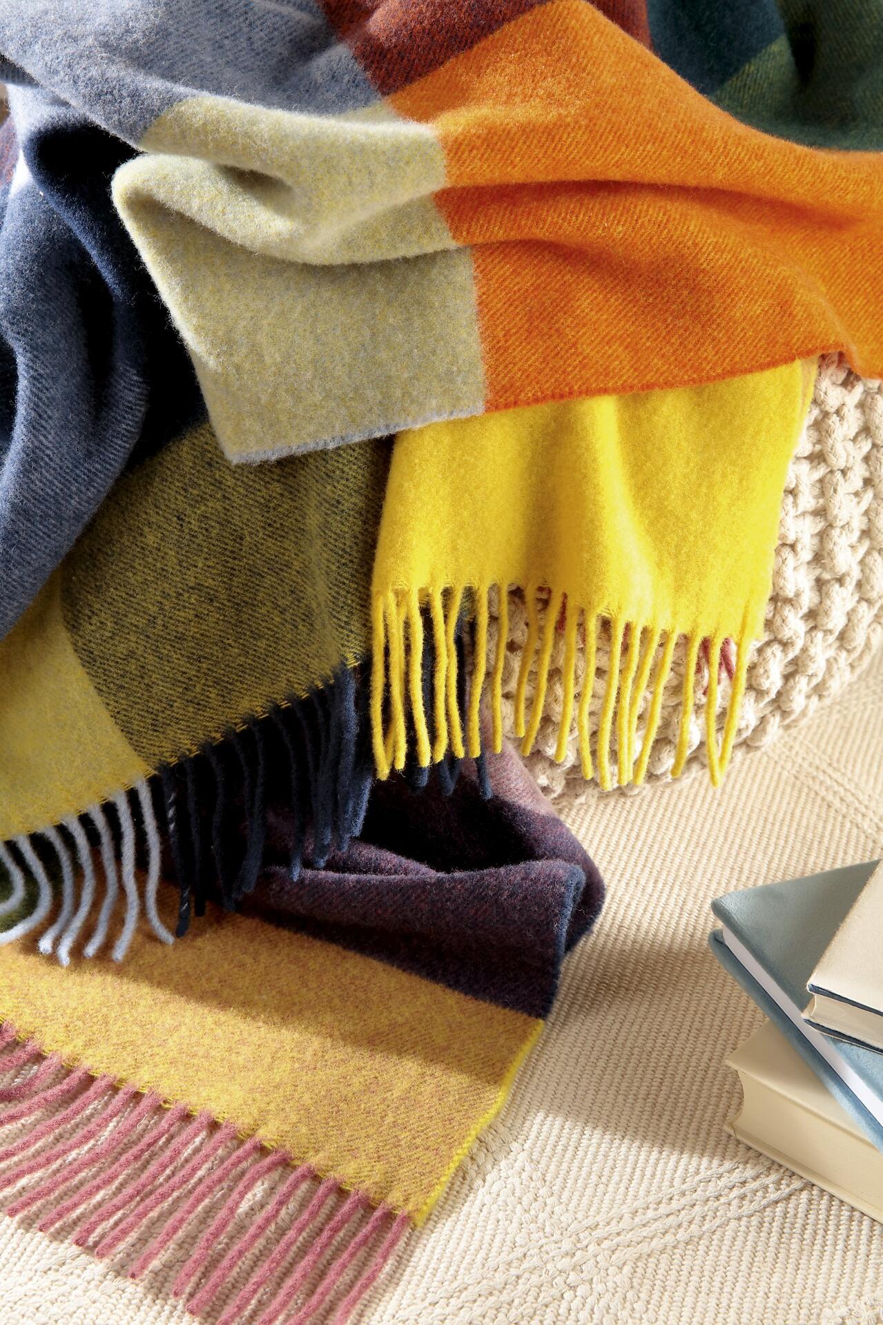Plaid Decke in warmen Farben mit Farbverlauf von Gelb über Orange bis Lila, lässig über einem Hocker drapiert, zeigt die grobe Strickstruktur und fransigen Kanten.