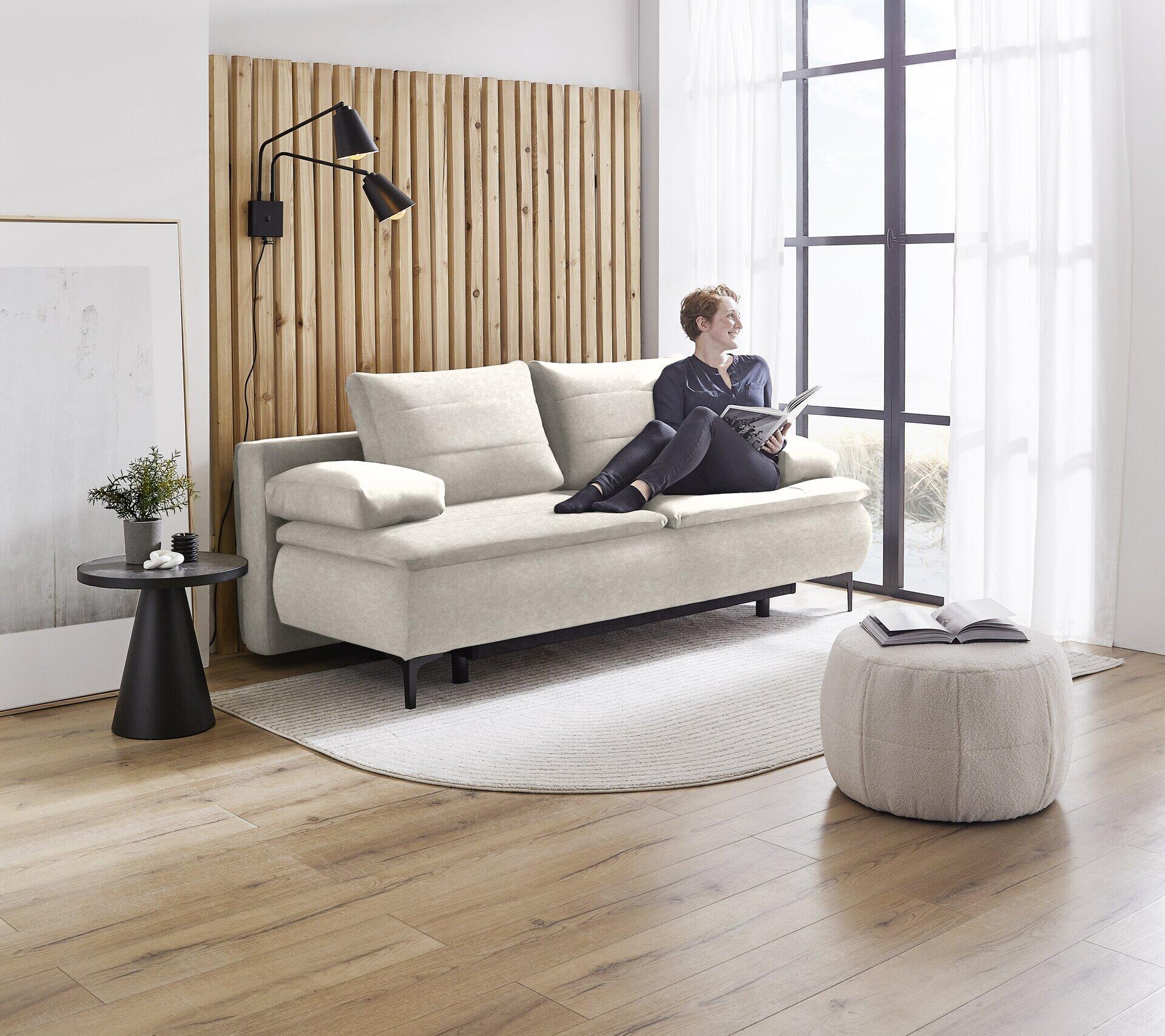 Melly Boxspringsofa im hellen Grau-Beige mit ausgezogener Schlaffunktion und Bettkasten, leicht erhöht fotografiert mit Person, Beistelltisch und Fußhocker.