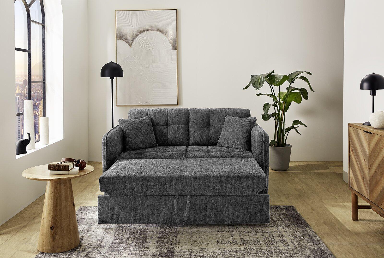 Schlafsofa Leon in grauer Chenille-Ausführung mit hoher Rückenlehne und großzügiger Liegefläche, die sich zur Schlaffunktion ausziehen lässt, dargestellt in einer stilvollen Wohnsituation mit Holztisch und Stadtblick im Hintergrund.