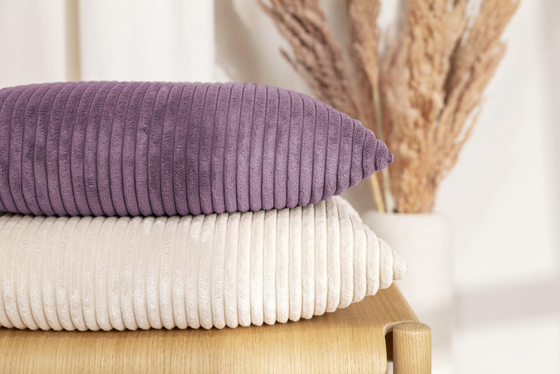 Violette Kissenhülle Shara aus Flauschcord mit strukturierter Oberfläche, dargestellt auf einem Holzmöbelstück zusammen mit einer beigefarbenen Kissenhülle.