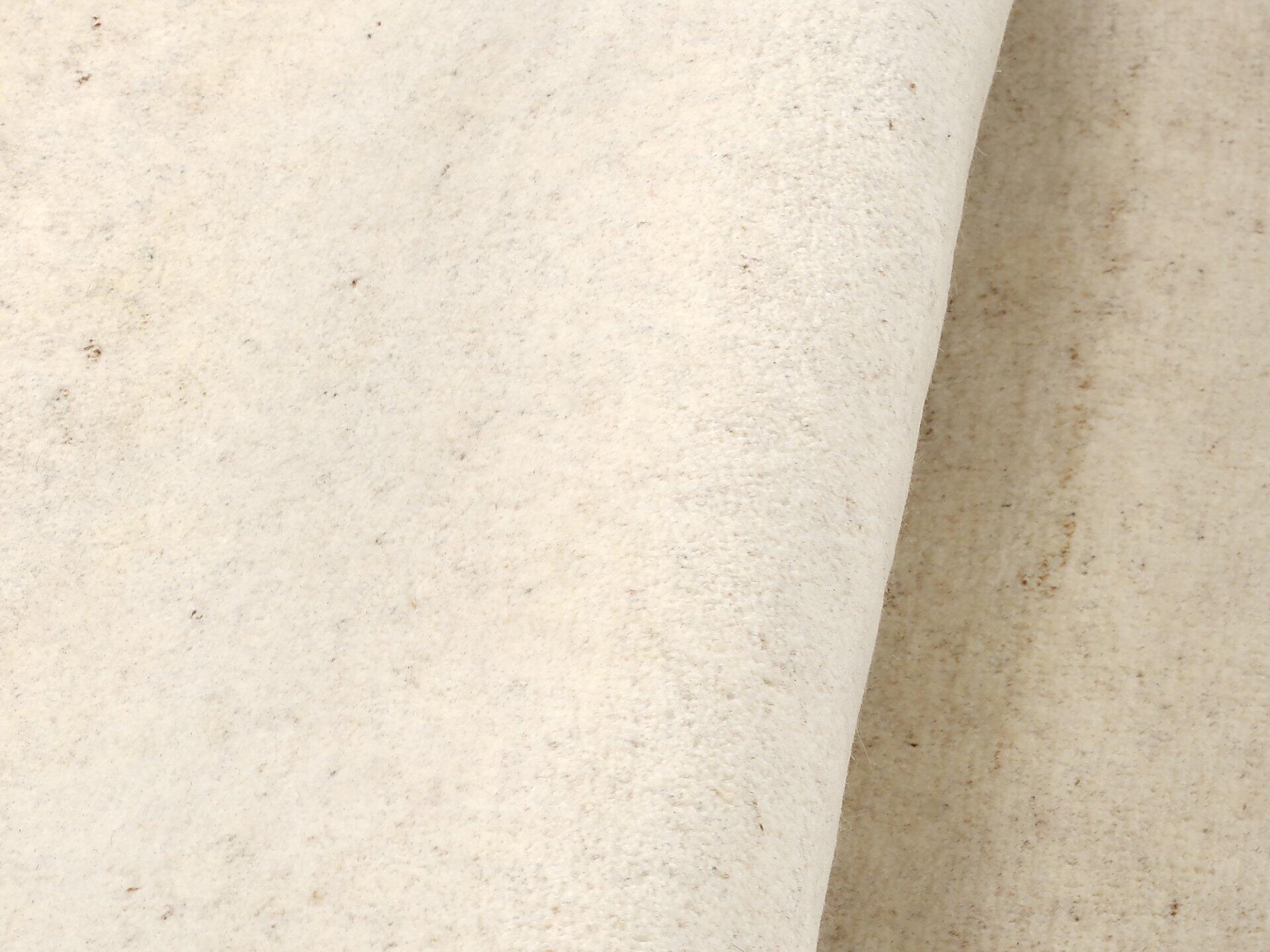 Detailaufnahme des cremefarbenen, samtigen Wollteppichs Assos mit feiner Struktur und dunkleren Pigmentierungen, die seine natürliche Optik unterstreichen.