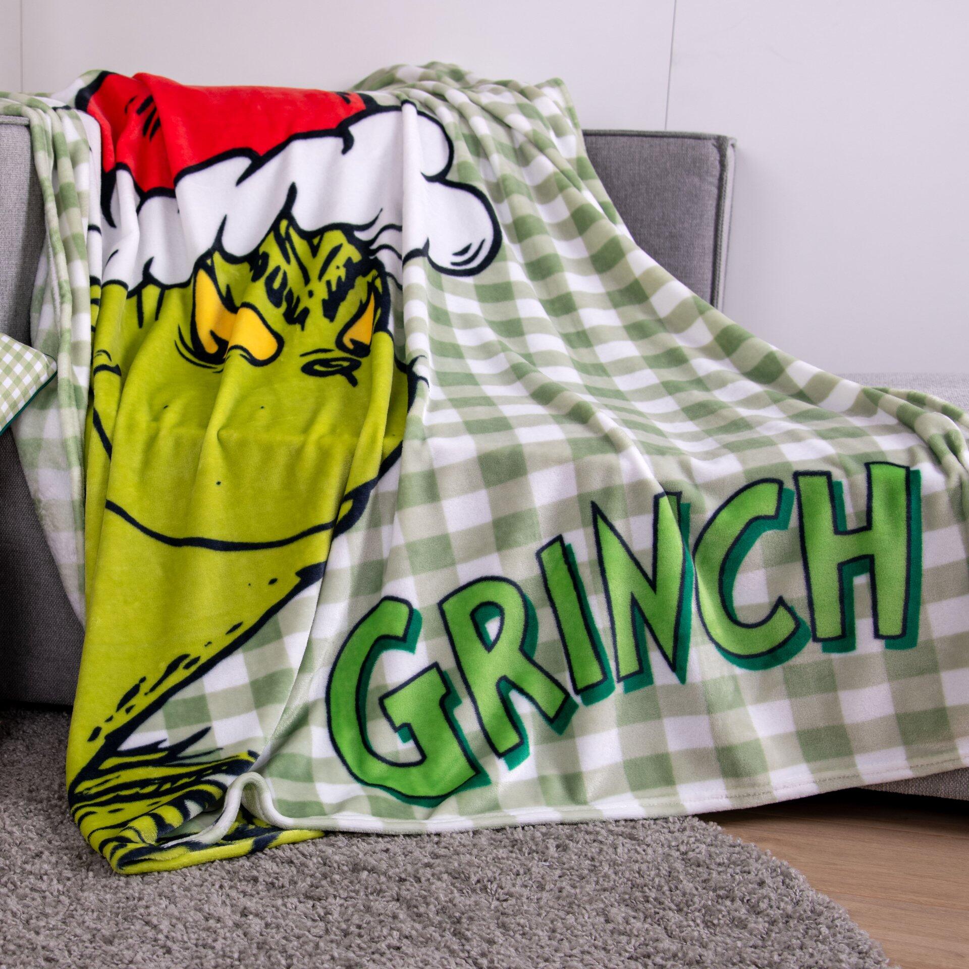 Kuschelige Decke "Grinch" aus 100% Polyester Wellsoft mit Motiv des Grinch und dem Schriftzug "GRINCH", abgebildet auf einer grauen Couch aus schräger, oberer Perspektive.