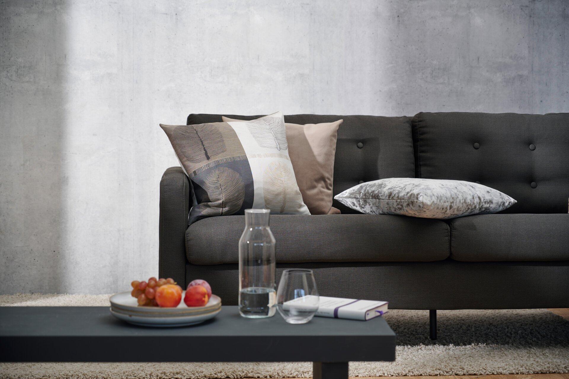 Graues Sofa mit dekorativen Kissen im Blattmuster und Reißverschluss, aus einer leicht erhöhten Perspektive fotografiert, zeigt den mittleren Bereich des Sofas und einen darunterliegenden Couchtisch.