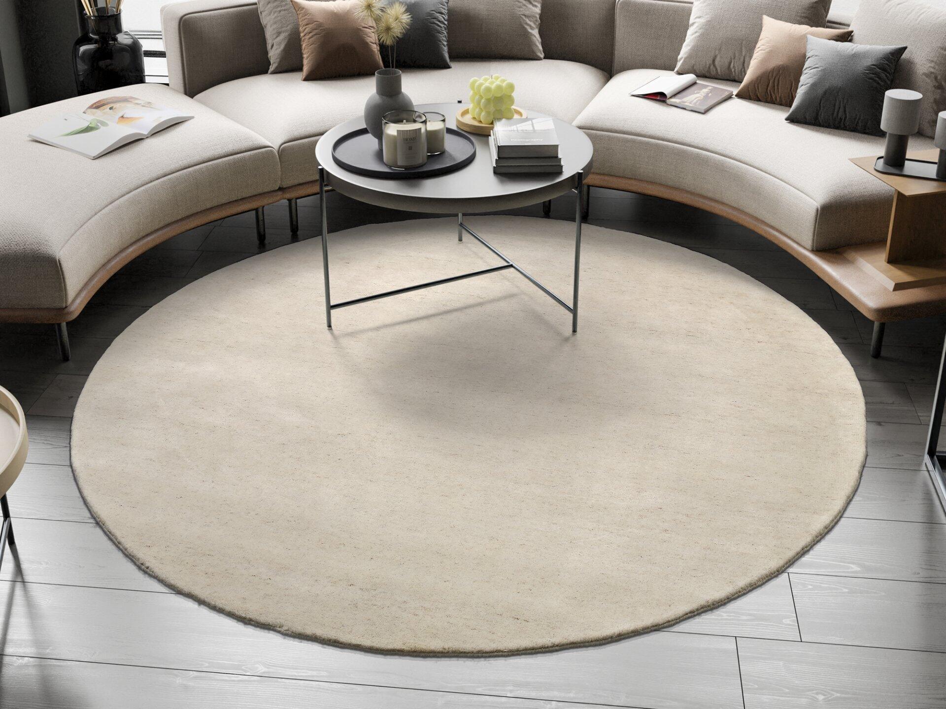 Detailaufnahme eines handgeknüpften Wollteppichs Modell Assos mit rundem Design und hellbeigem Farbton auf einem Holzfußboden, arrangiert mit einem Beistelltisch und Dekorationsgegenständen.