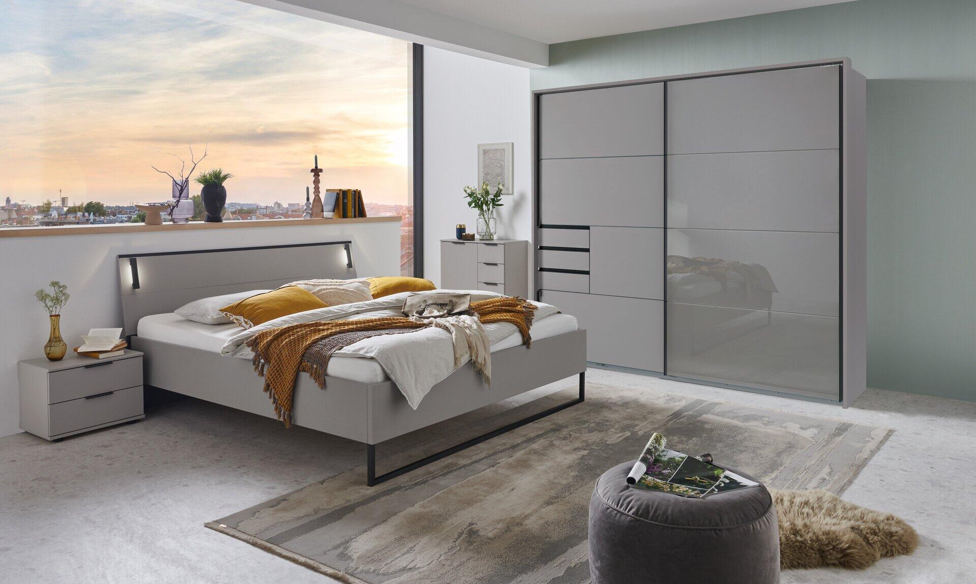 Weißer Schwebetürenschrank Tilburg mit glänzenden Türen und ausziehbarer Schublade, eingebettet in eine moderne Schlafzimmergestaltung mit grau lackiertem Bett und Nachttisch.