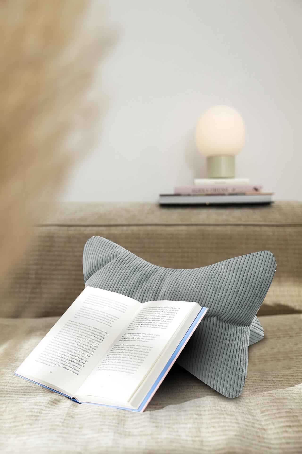 Leseknochen Inga: Graublaues Lesekissen mit Cordstruktur und Buchablage auf einem Sofa.