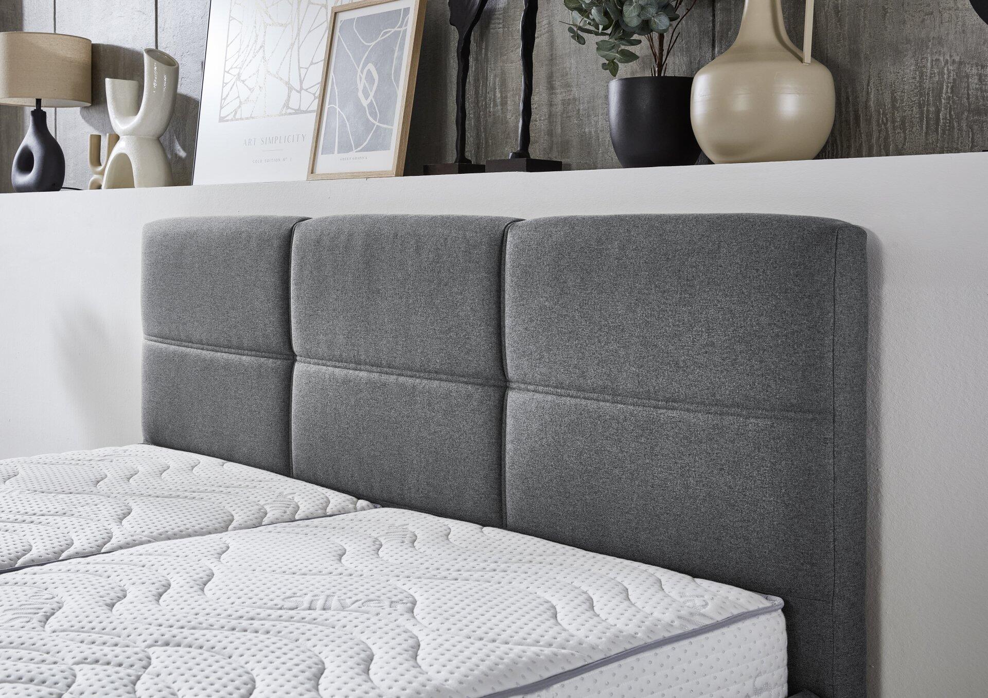 Graues Polsterbett Durango PB100 mit hochbasiertem Kopfteil, gestaltet mit strukturiertem Stoff und rechteckigen Kissenformungen, aus leicht unterer Perspektive aufgenommen.