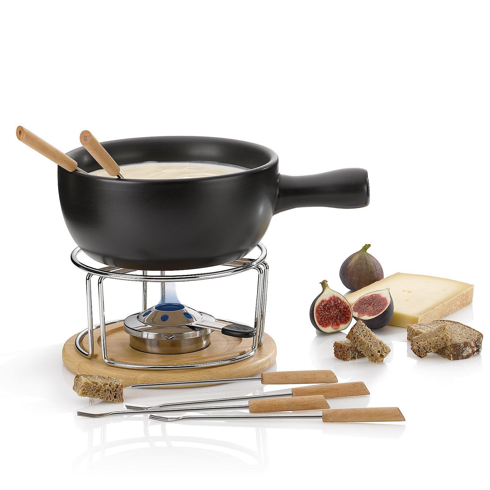 Käsefondue-Set Natura mit schwarzer Kasserolle, Gaskocher, Bambusplatte und Holzspießen, präsentiert mit Käse und Beilagen auf reflektierender Oberfläche.