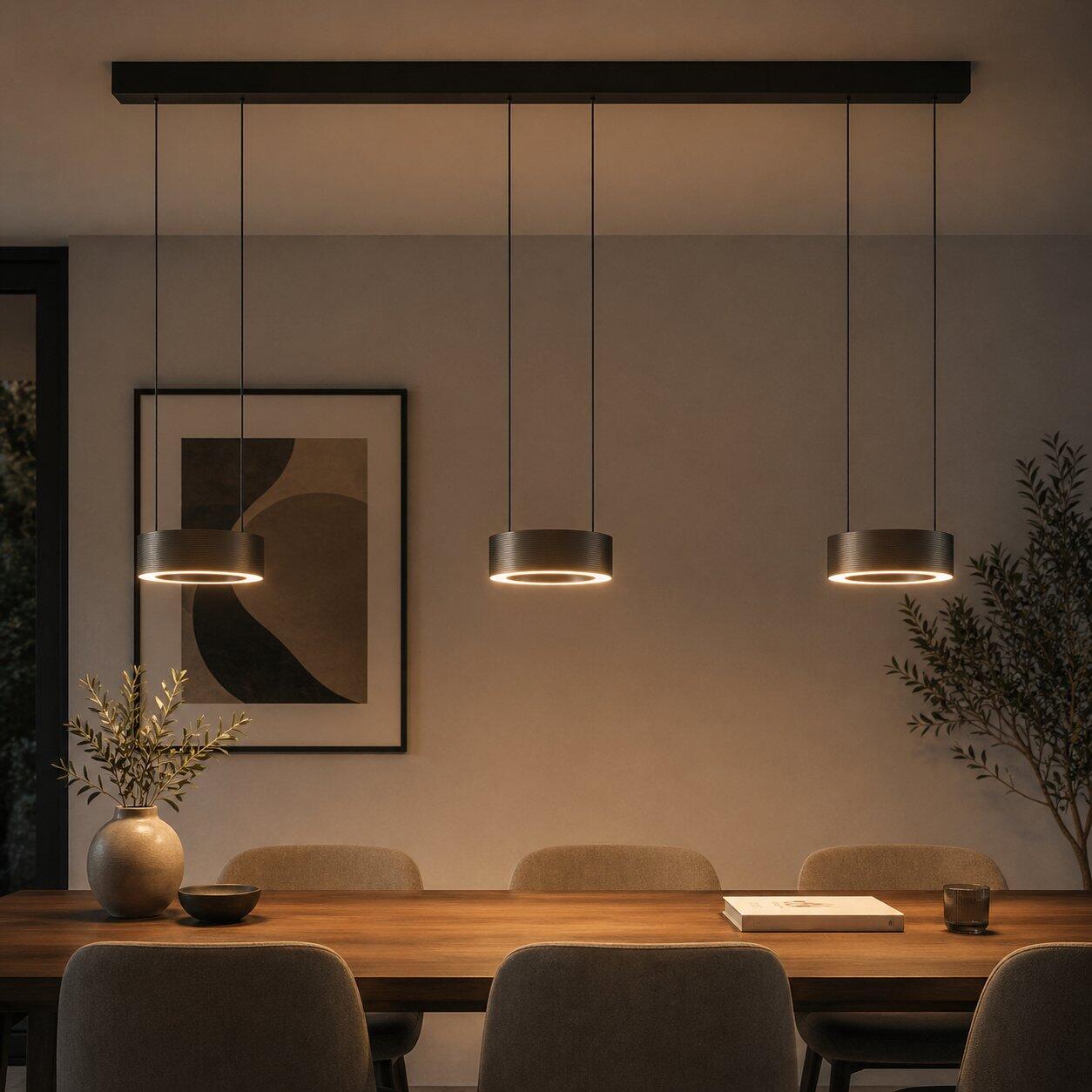 Pendelleuchte Axion mit zylindrischem Design, die über einem Holztisch eine warme, gelbliche Beleuchtung erzeugt und dessen Funktion als Essplatz unterstreicht.