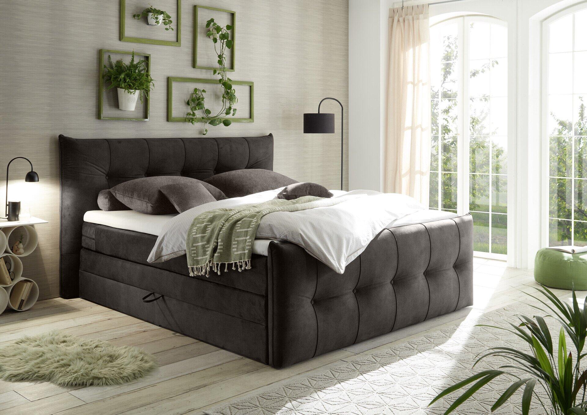 Modernes Boxspringbett Maracana 6 in Grau mit gestepptem Kopfteil, Tagesdecke und Kissen auf dem Bett, abgebildet aus leicht erhöhter Drei-Viertelperspektive.