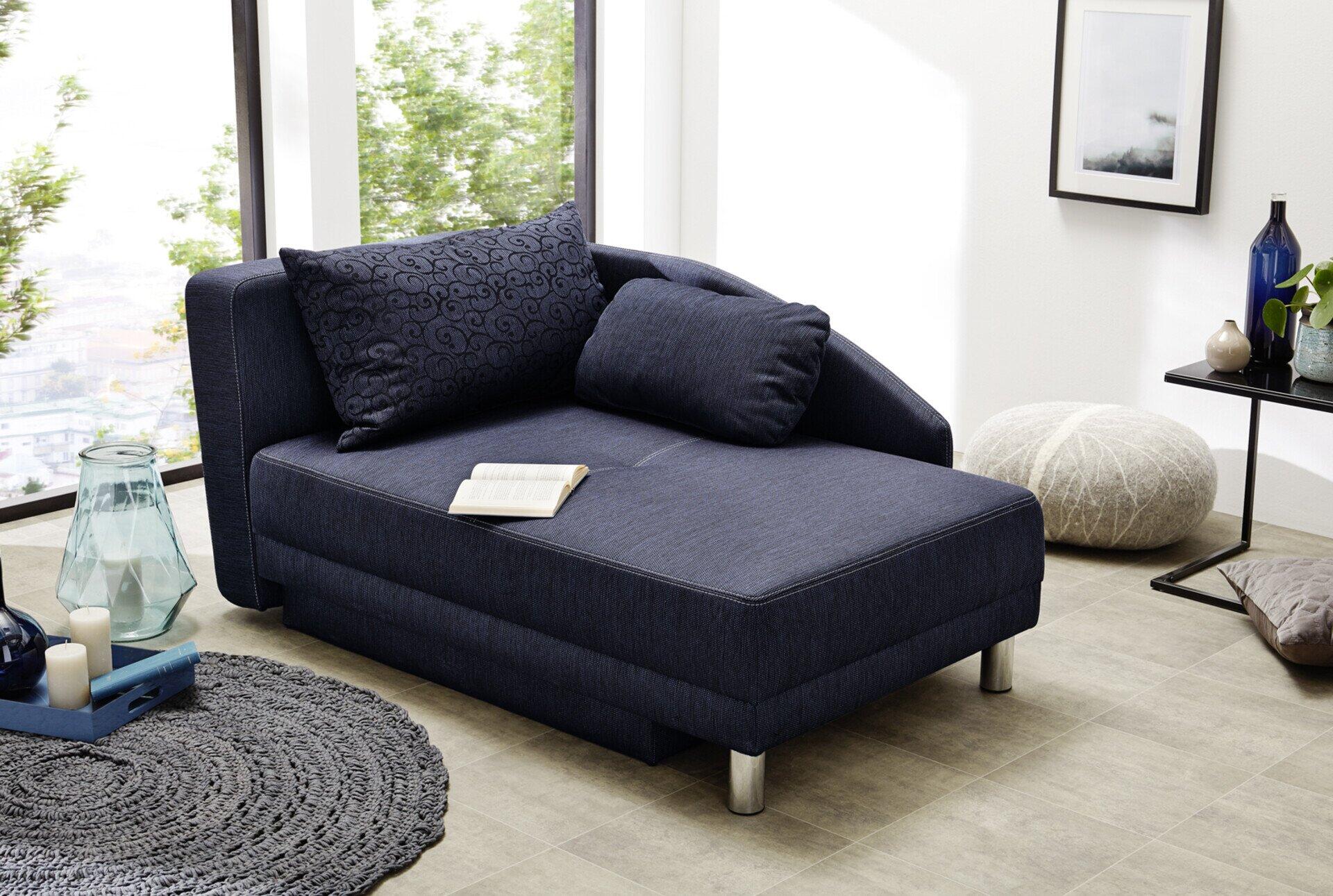 Dunkelblaues Schlafsofa "Roy" mit Ottomane, chromfarbenen Füßen und hoher Rückenlehne, detailliert mit Buch und Kissen auf der tiefen Sitzfläche.