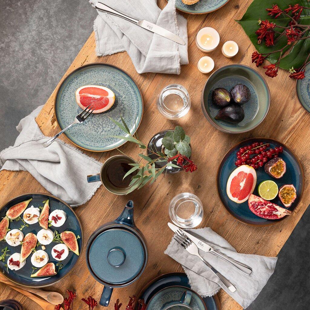 Draufsicht eines gedeckten Tisches mit Lebensmitteln und Küchenutensilien, im Mittelpunkt eine dunkelblaue Pfanne mit blauem Griff, umgeben von Tellern mit Früchten und Salat.