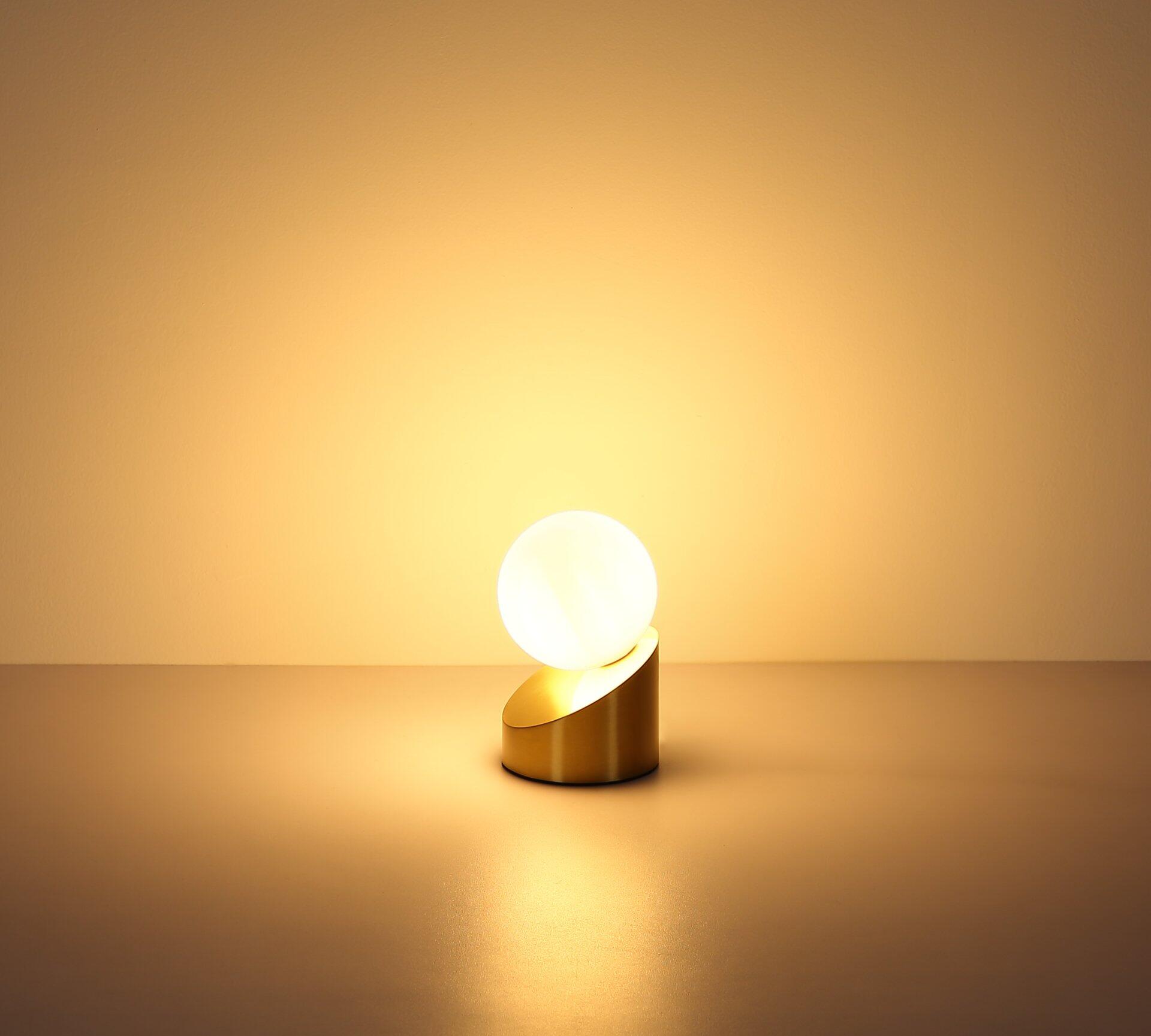 Tischleuchte ALISA mit runder Glaskugel und goldener Halterung, beleuchtet mit warmweißem Licht, auf goldenem Untergrund.