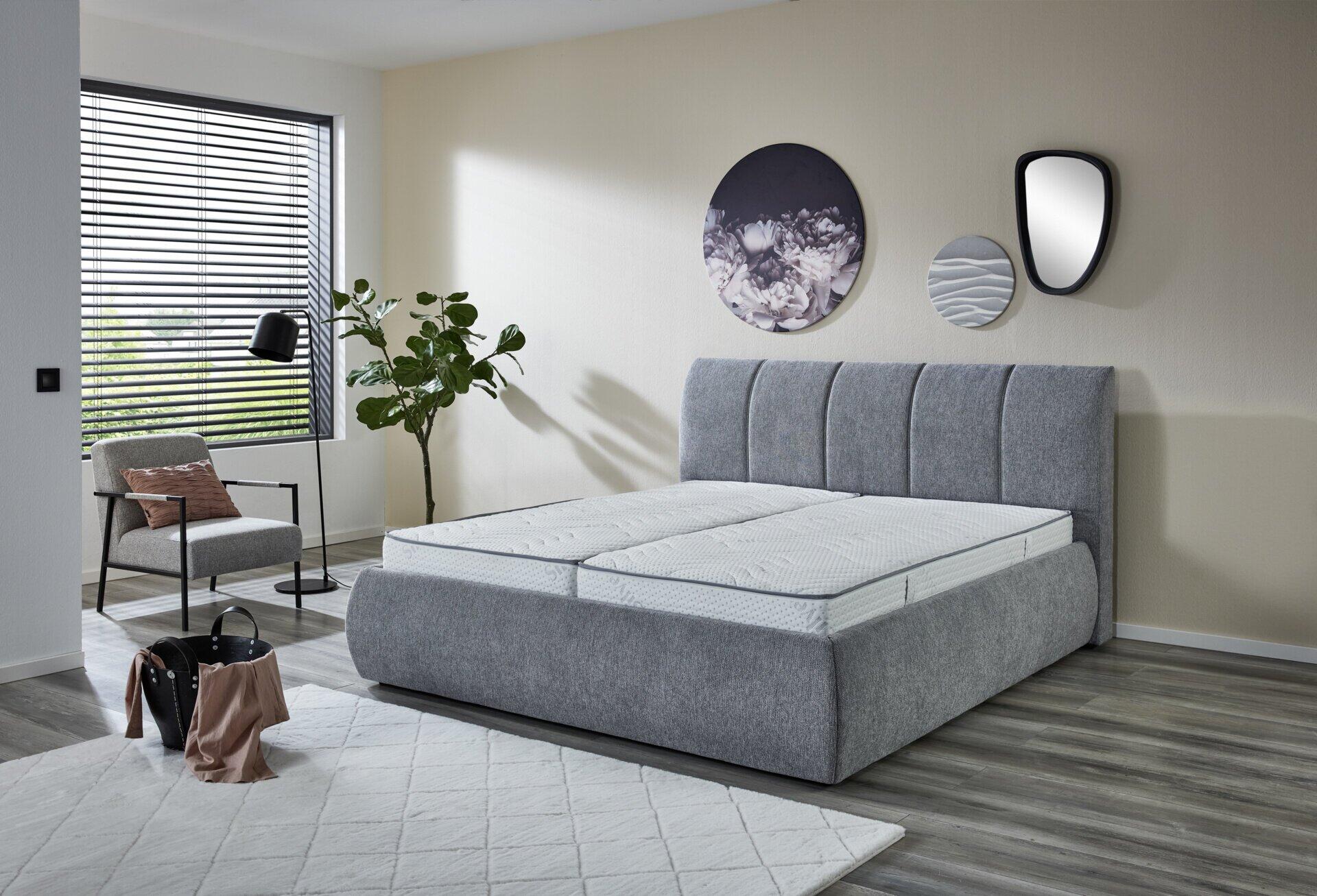 Polsterbett Vail PB130 mit grauem Stoffbezug und vertikaler Steppung, inklusive integriertem Bettkasten, präsentiert in einem hellen Schlafzimmer.
