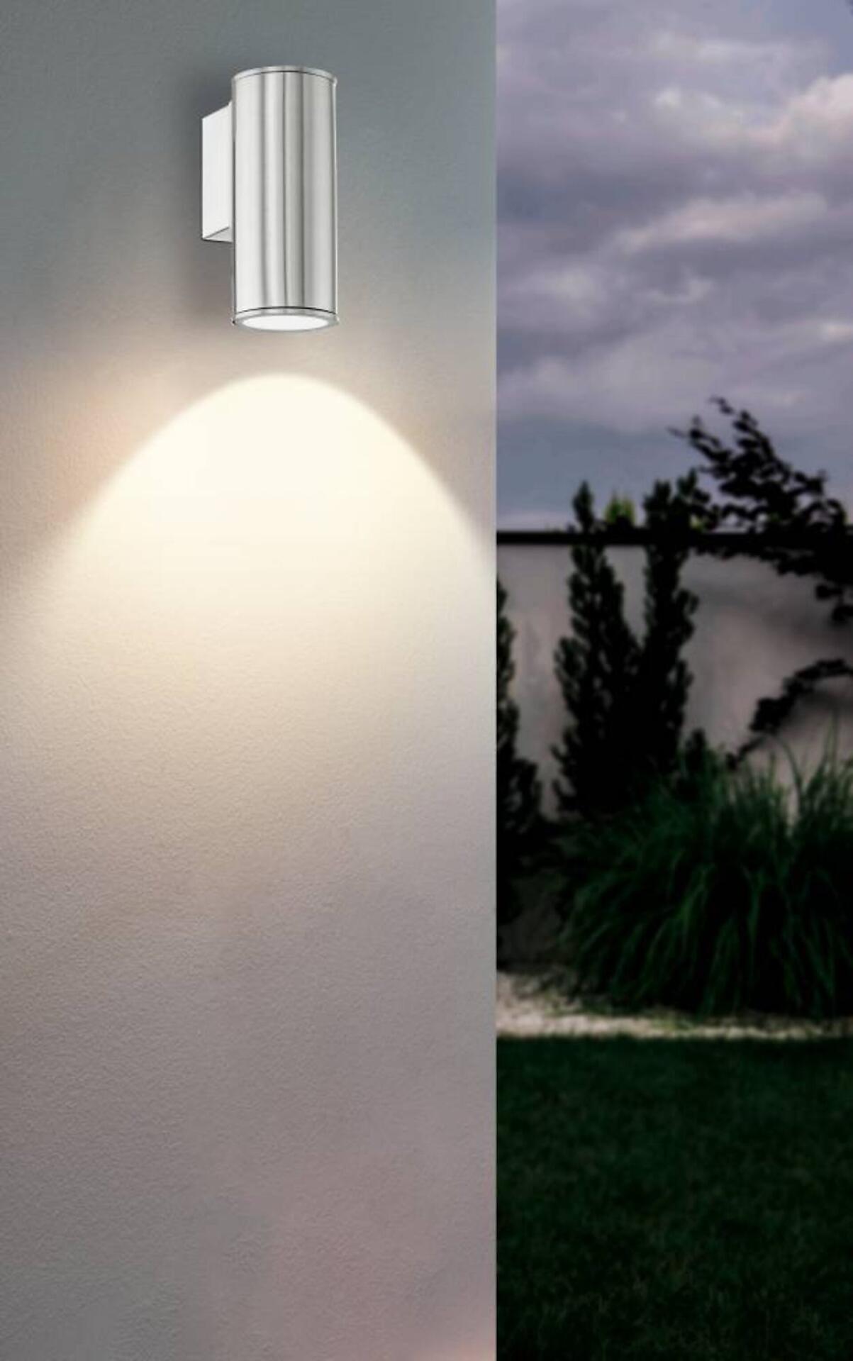 Silberfarbene Zylindermantel-Wandleuchte wirft warmweißes Licht auf eine grüne Landschaft.