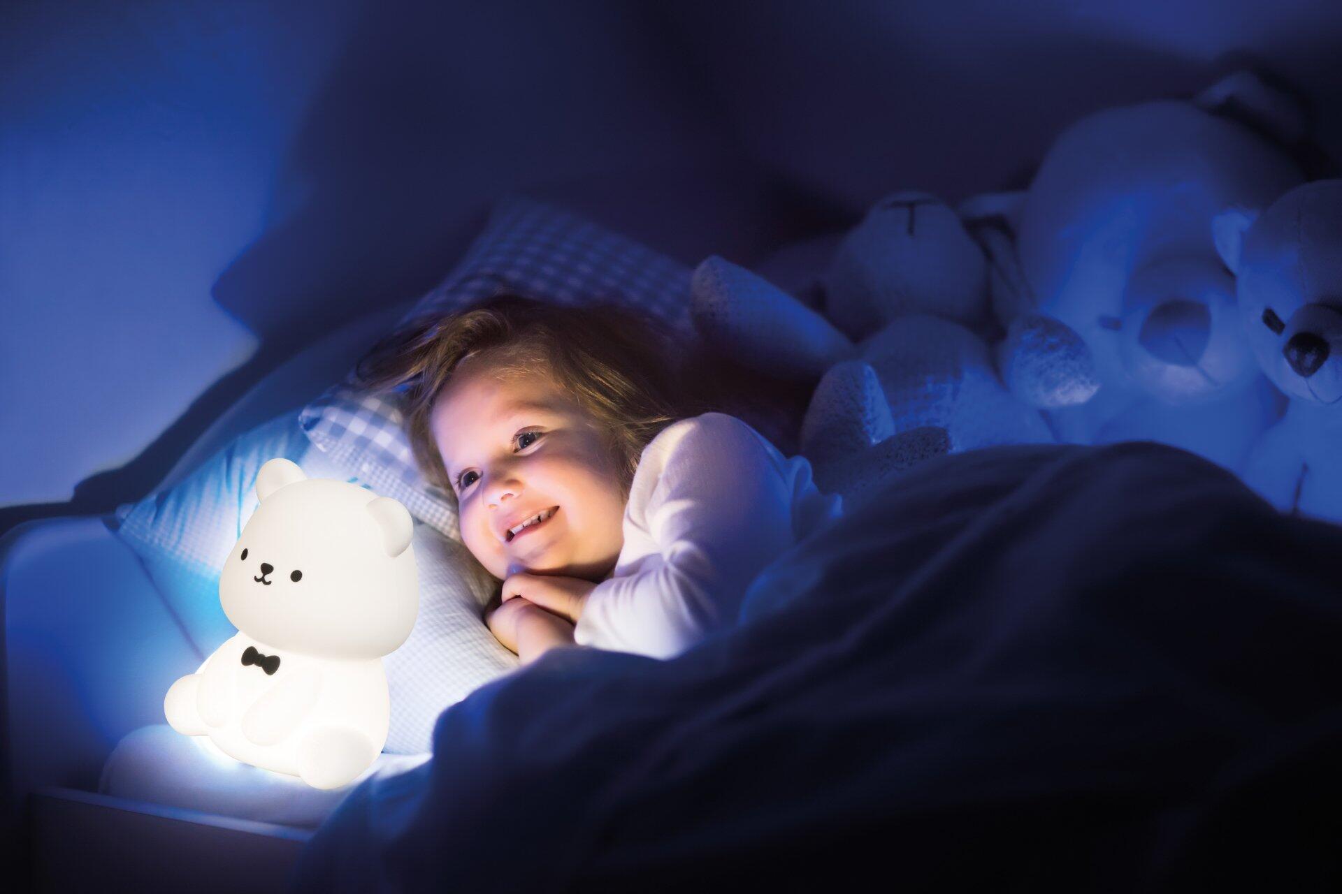 Nahaufnahme einer weißen, rundlichen Tischleuchte, Karl, die neben einem Kind im Bett sanftes Licht ausstrahlt und als Nachttischlampe eine beruhigende Atmosphäre schafft.