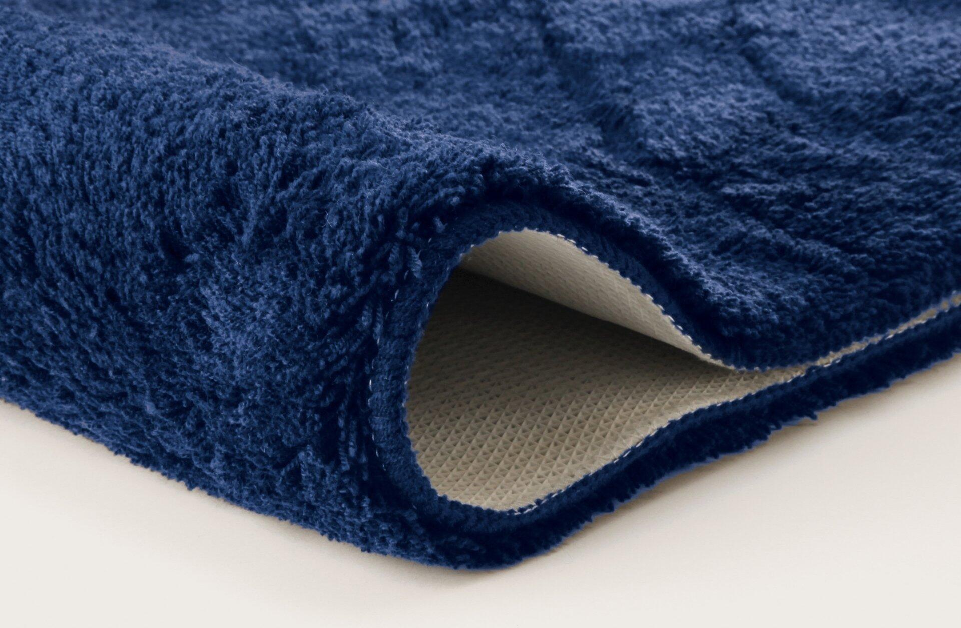 Saturn Badteppich in Marineblau: Detailaufnahme des weichen, flaumigen Teppichs mit hochgeklappter Kante, die das wabenförmige, rutschfeste Rückseitenmaterial zeigt.