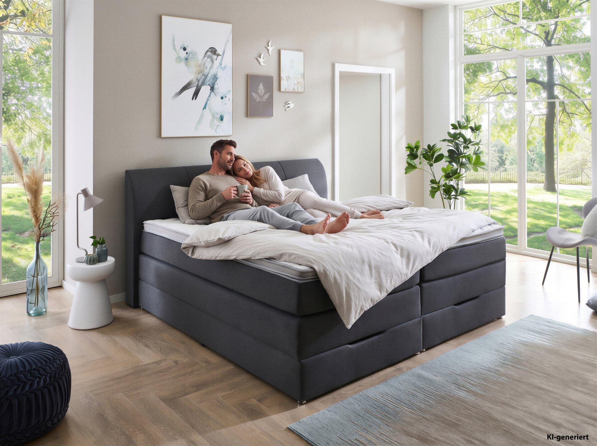 Boxspringbett Modesto BX1440 mit grauem, gepolsterten Kopfteil und integriertem Stauraum, dargestellt in einem hellen Schlafzimmer mit einem Paar, das entspannt auf dem Bett liegt.