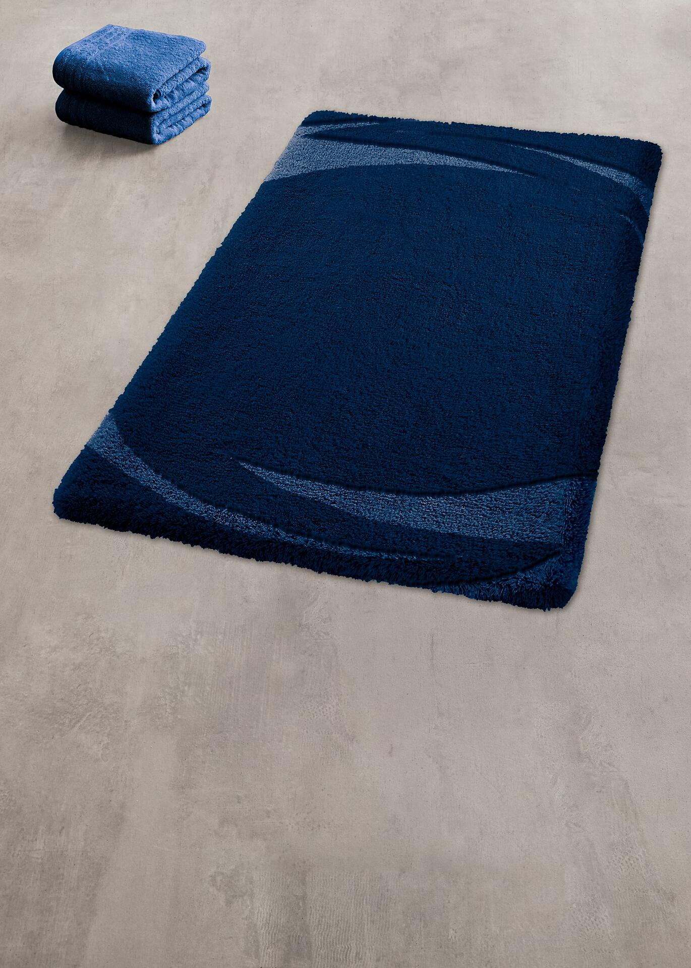 Dunkelblauer Badteppich Mystic mit strukturierter Oberfläche auf grauem Boden, daneben gefaltete Handtücher.