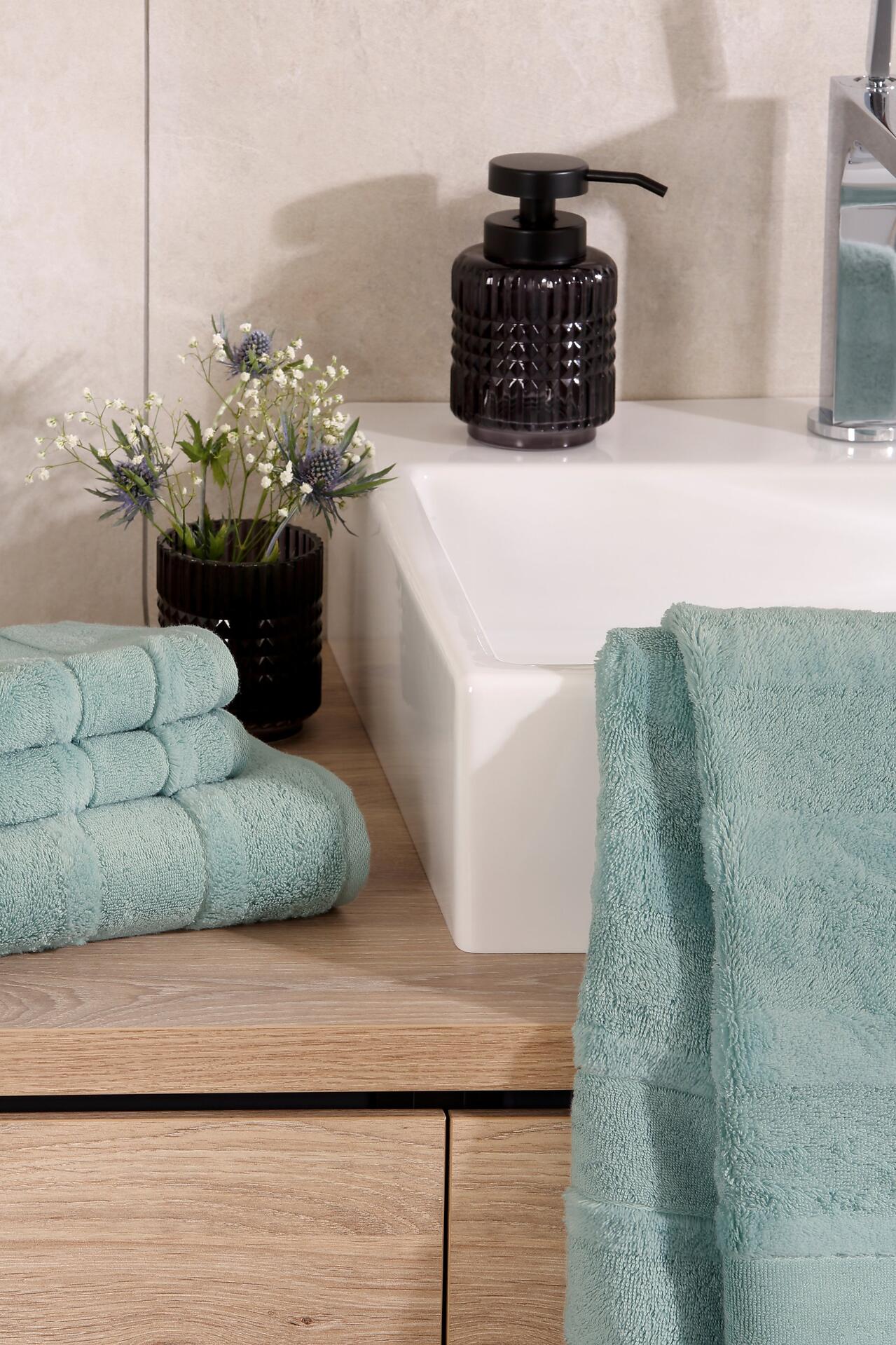 Flausches Duschtuch Noblesse² in Emerald/Teal-Farbton liegt auf einer Holzablage neben einem weißen Waschbecken.