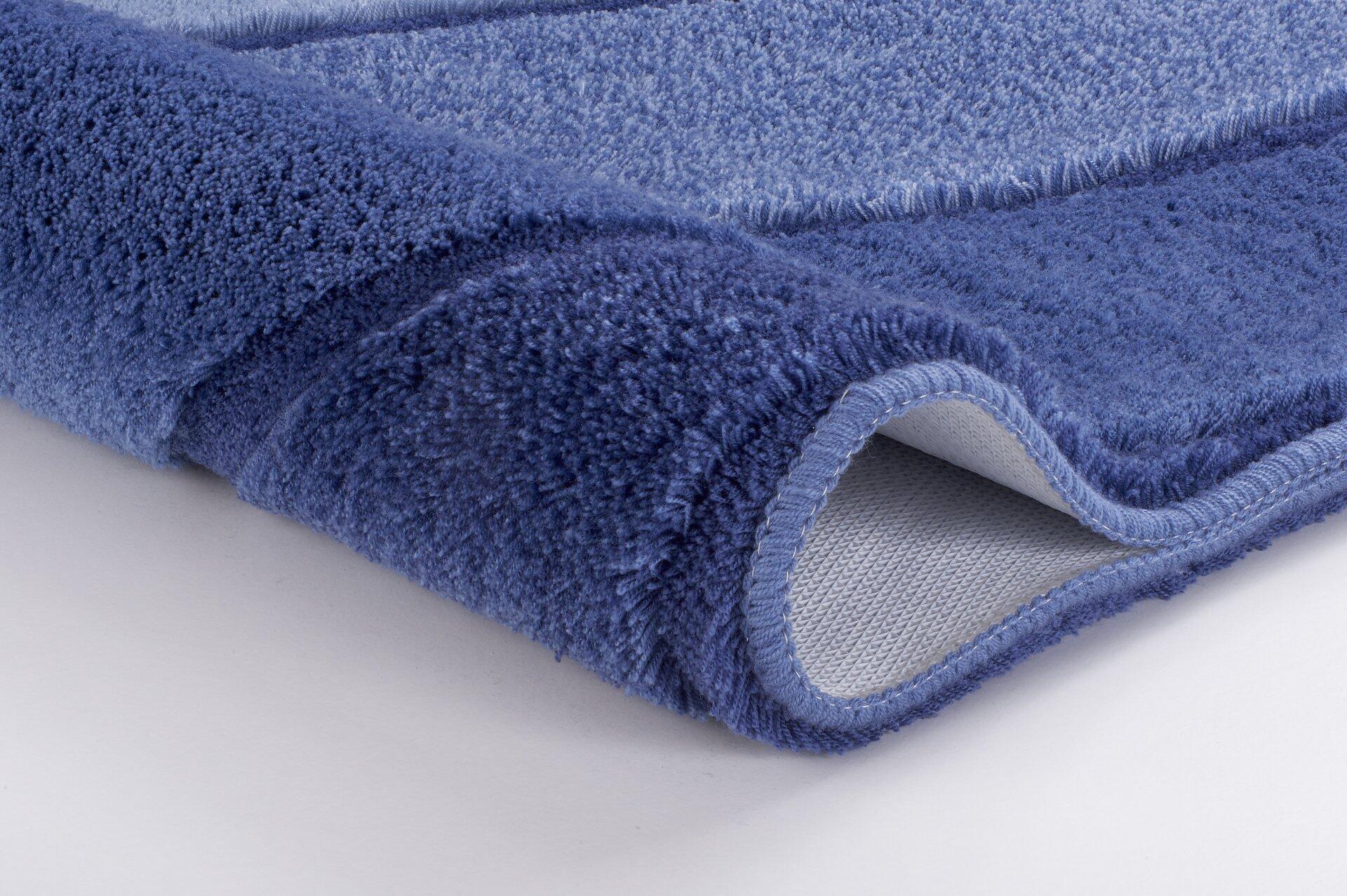 Gefalteter Badteppich Serenade in Iceblue mit flauschiger, strukturierter Oberfläche und hochgeklapptem Rand, der ein graues, rutschfestes Gewebe zeigt.